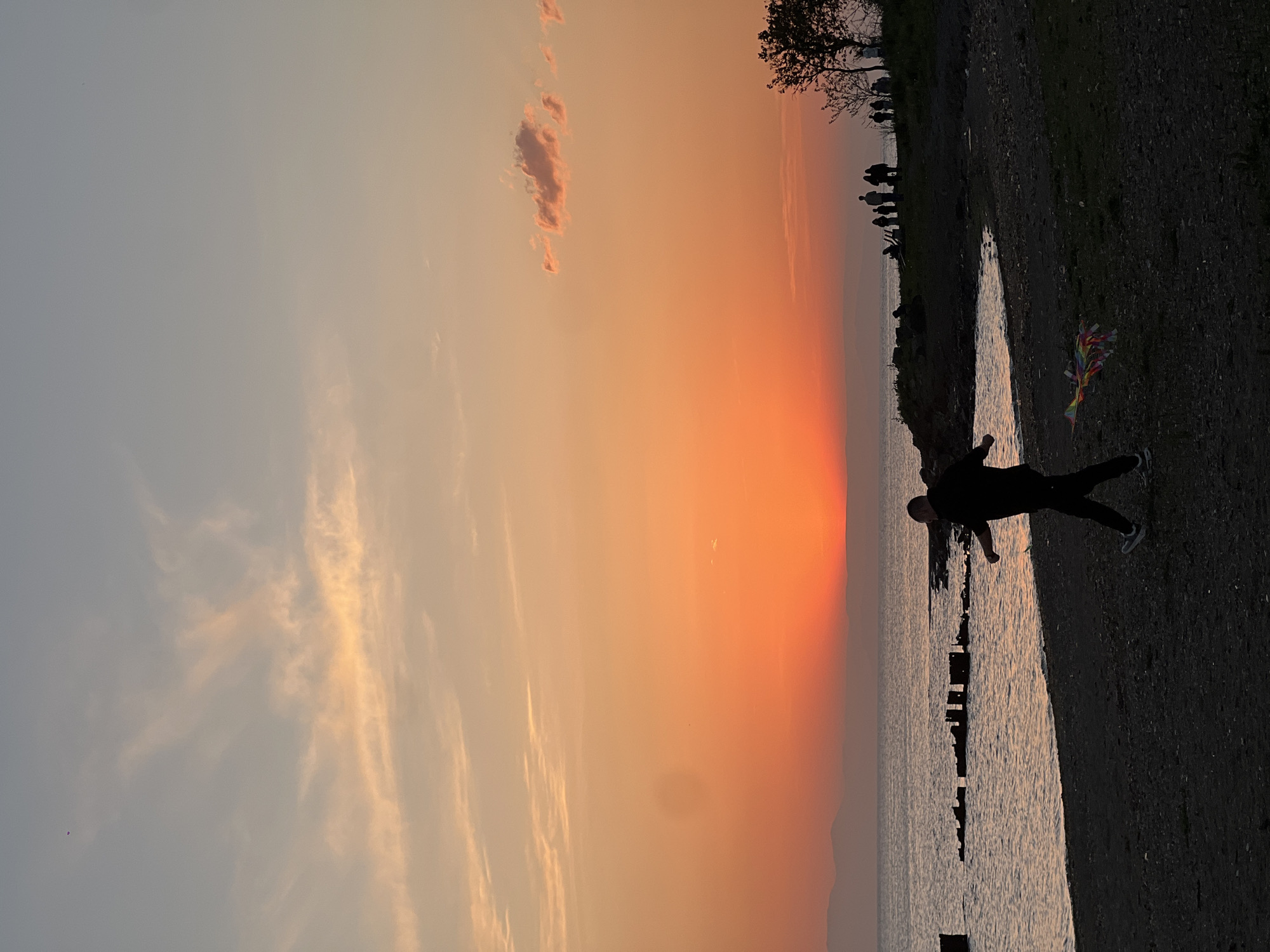 The image depicts a sunset scene in what appears to be Vladivostok, Russia. In the foreground, a person stands on a pebble beach near the shoreline. Their silhouette suggests a casual posture, possibly with outstretched arms. A colorful kite is discarded nearby on the ground. Along the shoreline, old wooden poles are embedded in the water. In the distance, other people appear to be gathered along the shoreline. The sky transitions from a bright orange near the horizon to a pale blue-grey higher up, with clouds scattered throughout. The horizon line shows indistinct landforms.