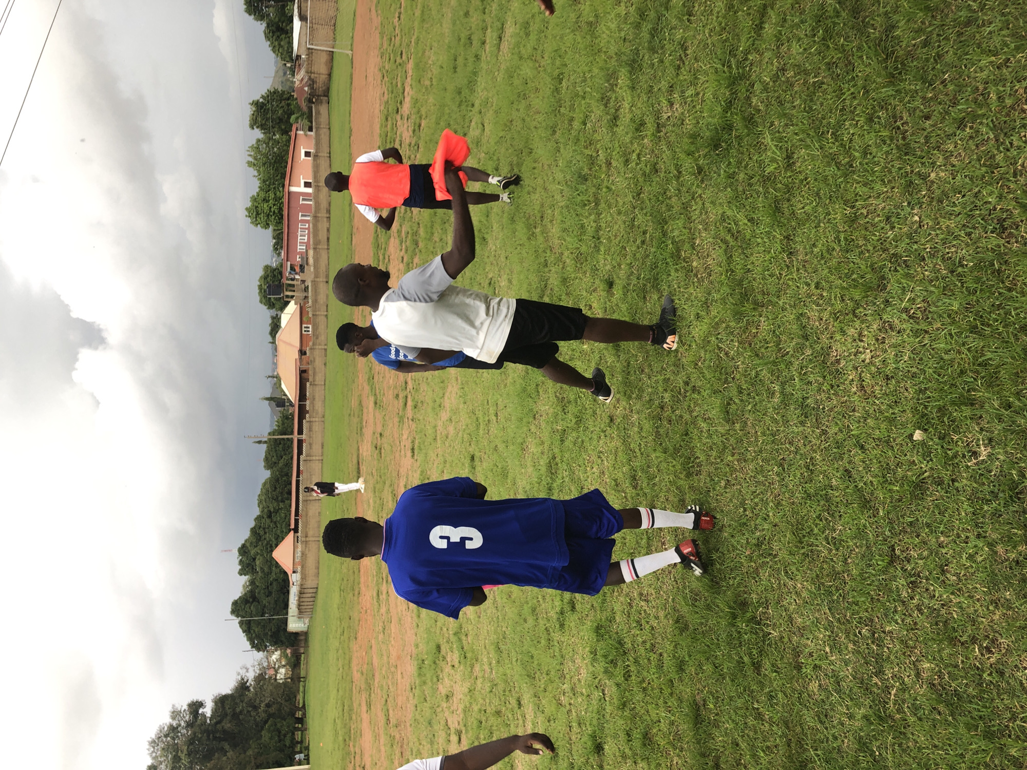 In the city of Jos, Nigeria, on a grassy sports field under a cloudy sky, four young Black men are engaged in a practice session. The man in the foreground, wearing a gray and white t-shirt and black shorts, holds a bright orange cloth in his right hand, appearing to be instructing or demonstrating. To his left, a man in a blue jersey with the number "3" on the back and matching blue shorts stands facing away from the camera, his white socks with red and black stripes visible. Behind these two, another man in a white t-shirt and black shorts is also facing away, wearing a red vest. Further in the background, a fourth individual in a white shirt and dark pants stands near a fence. The field is uneven, with patches of dirt and sparse grass, and a goalpost is visible in the distance. Residential buildings and trees are scattered around the perimeter of the field.