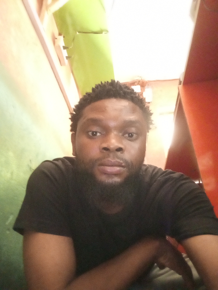 A single adult male is depicted from the chest up, centered in an indoor environment in Magboro, Nigeria. The individual has dark skin, a full black beard, a mustache, and short, dark, textured hair. He wears a plain black short-sleeved t-shirt. His expression is neutral, and he is looking directly at the camera. The background consists of a light green wall on the left and a reddish-orange wall on the right. In the upper left background, a section of light green, possibly fabric or plastic, is visible, appearing folded or crumpled. Below this on the light green wall, a white electrical outlet or switch plate is present. The scene is brightly lit, with strong illumination originating from an unseen source above.