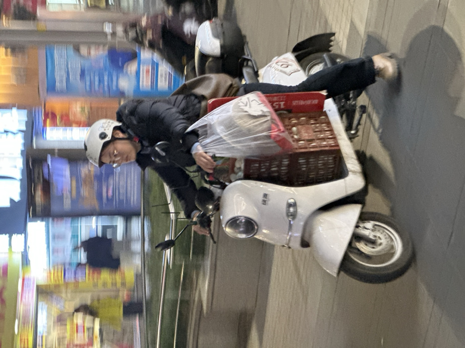 The image depicts an urban street scene at night in Jiangjin, China. A person, appearing to be an adult, is captured in motion while riding a white electric scooter on a paved surface. The rider wears a white helmet with a visible chin strap, glasses, a dark puffer jacket, dark trousers, and light-colored boots. Their hands grip the handlebars.
The electric scooter features a single front headlight, rearview mirrors, and a white storage box mounted on the rear rack. A large, brown, perforated plastic crate is secured in the scooter's footwell, containing various items. A transparent plastic bag filled with contents hangs from the handlebars, draped over the crate. A red rectangular object is wedged vertically between the crate and the scooter's body on the right side. Motion blur around the scooter's wheels and in the background indicates movement.
In the background, illuminated commercial storefronts with Chinese characters are visible. Blurred figures of other individuals are present near these establishments, including one person wearing a yellow jacket. A low concrete barrier topped with a silver metal railing separates the foreground area from the background storefronts, with some greenery visible atop the barrier. Another blurred vehicle is partially visible behind the main scooter on the right side of the frame.