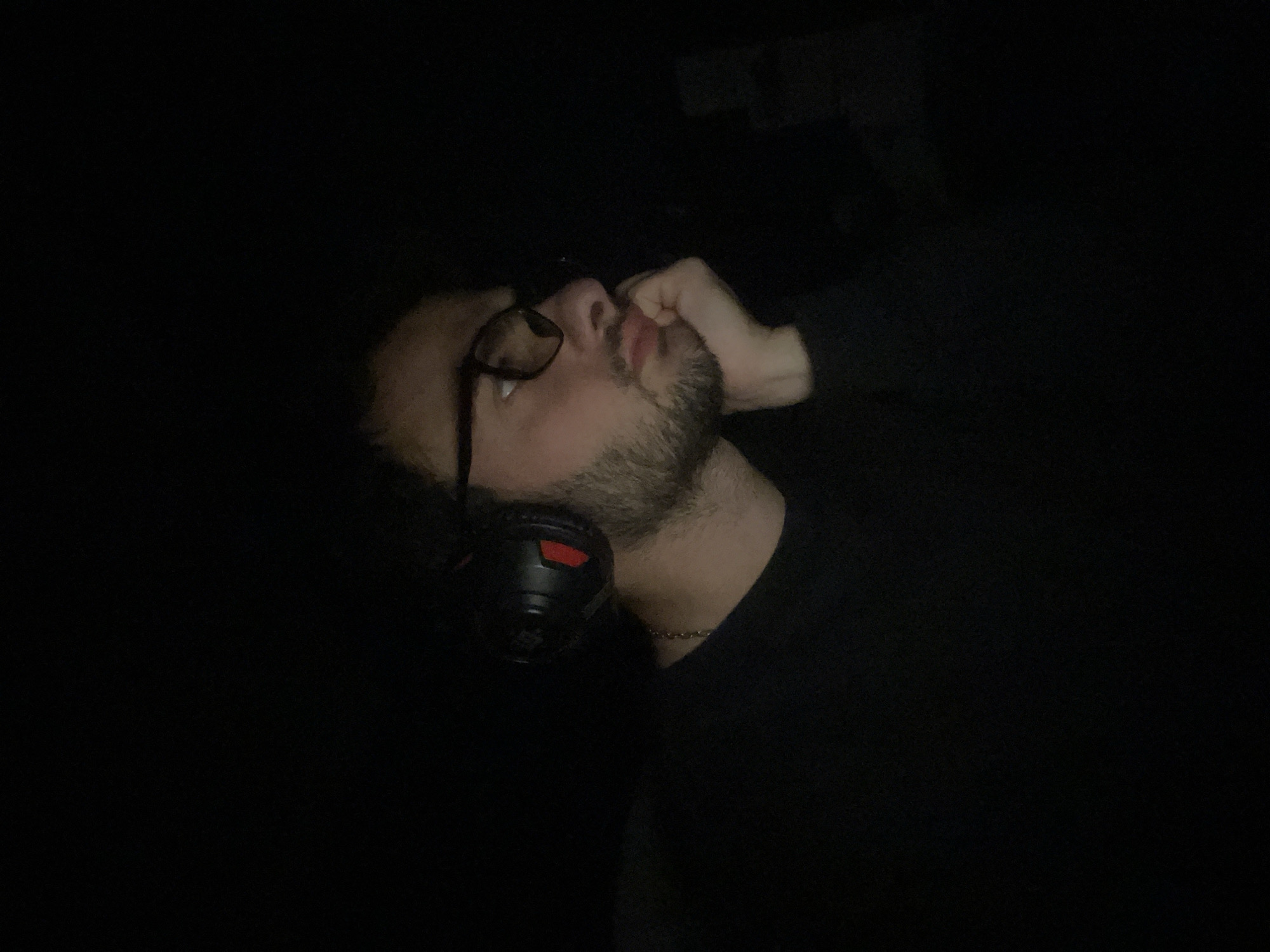 A person with dark hair and a beard is depicted in near darkness, wearing glasses and a headset. The person's fist is near their mouth. The environment is almost entirely black. The city of Tbilisi, Georgia, is not determinable from the visual information provided.