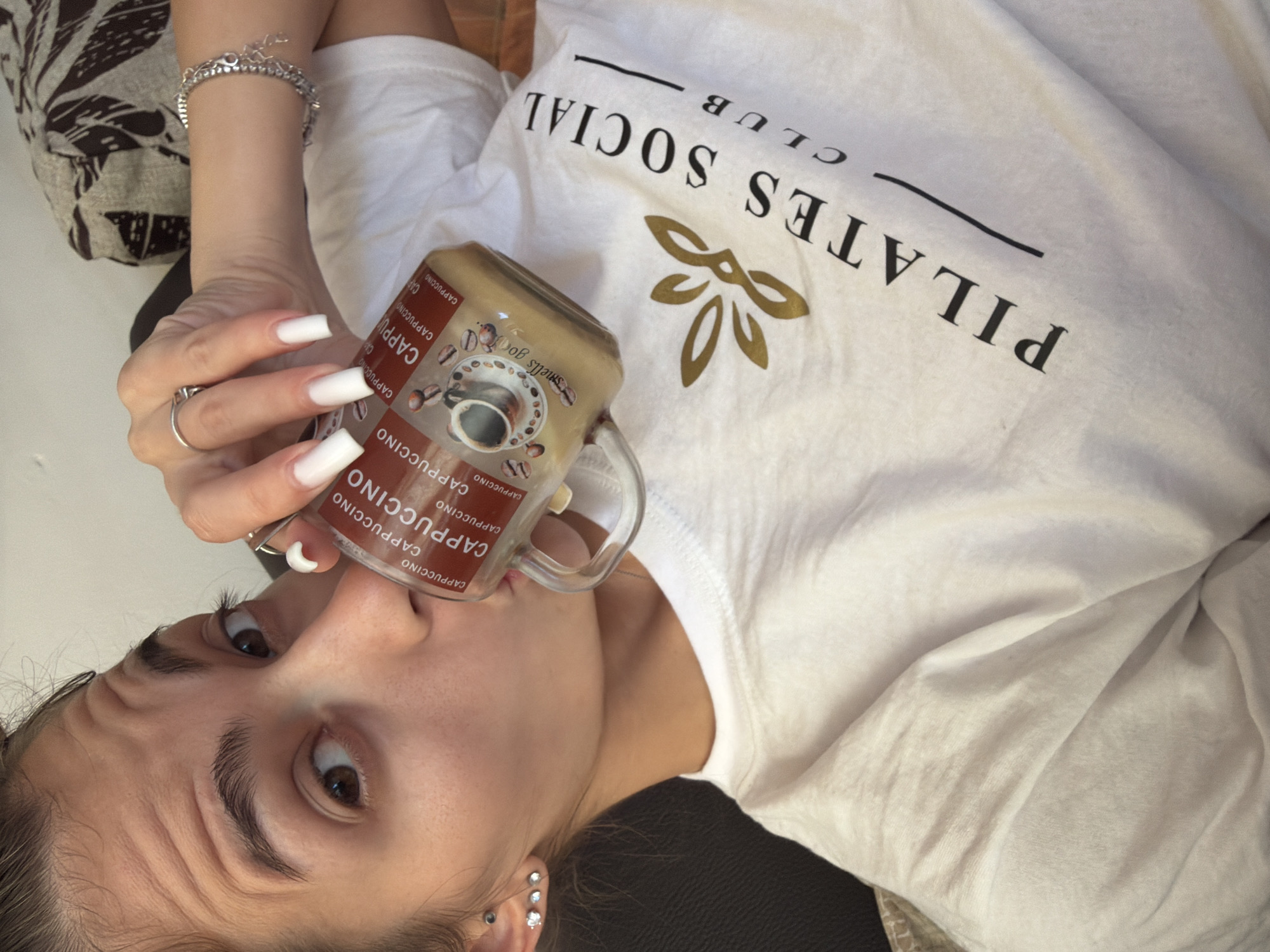 A young woman with long white acrylic nails and a silver ring on her right index finger is drinking from a glass mug with "Cappuccino" printed on it. She is wearing a white t-shirt with "PILATES SOCIAL CLUB" printed on the front. The woman has brown eyes and is looking at the camera. She is also wearing stud earrings in her left ear. The background is out of focus, but appears to be a living room with a sofa.