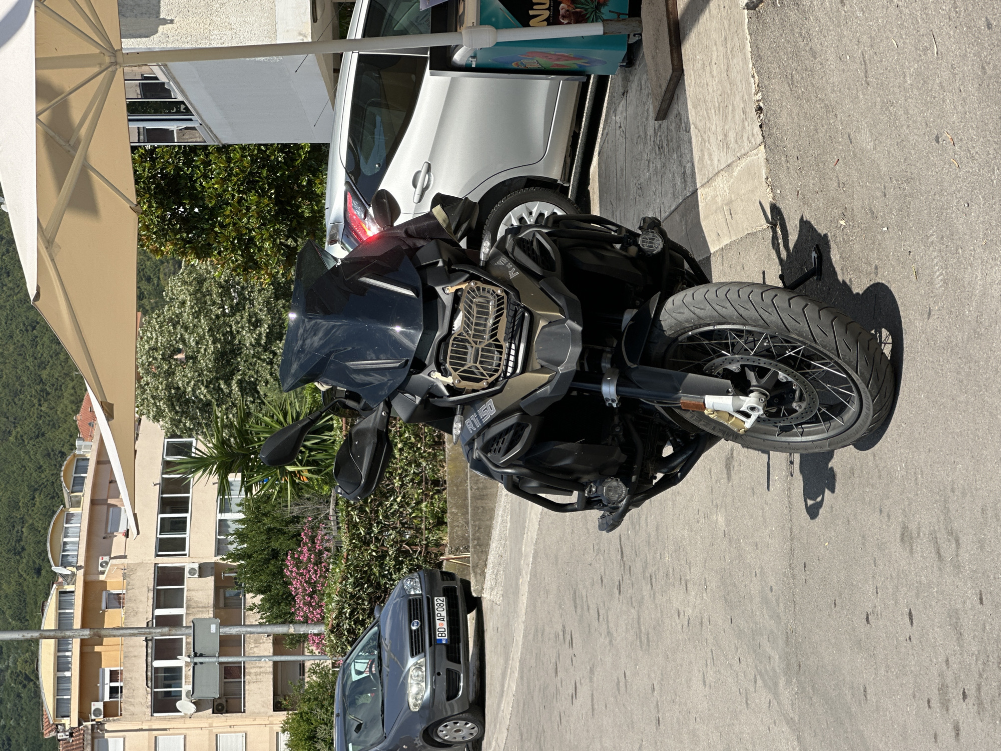 An outdoor, eye-level, medium shot shows a black BMW R 1250 RT motorcycle parked on an asphalt road. The motorcycle faces the camera. The motorcycle has a dark windshield, silver accents on the engine and a silver luggage rack. The front wheel is turned slightly to the left. A grey Fiat Punto is parked in the background to the left of the motorcycle, and a white car is parked behind the motorcycle to the right. A light-colored umbrella shades part of the scene. Buildings with balconies and trees are visible behind the cars. The location is Budva, Montenegro.