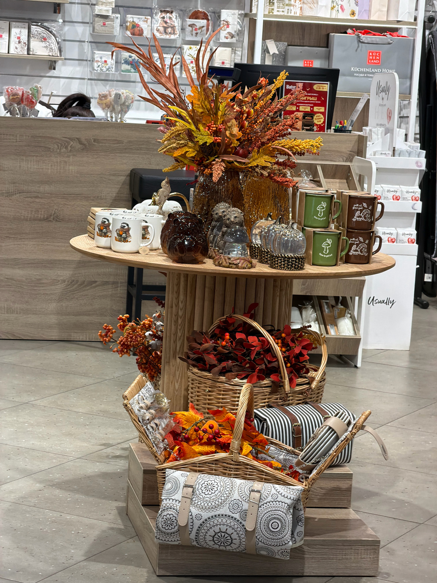 The image displays a retail display, likely in a home goods or gift shop. The foreground features a tiered display of baskets, with a woven basket containing autumn-themed decorations and a striped picnic bag. Another basket below holds a rolled-up blanket with a mandala pattern. The centerpiece is a round table showcasing mugs, decorative owl figures, and glass pumpkins. A large autumnal floral arrangement dominates the top of the display. In the background, shelves are stocked with various items, including packaged goods and shopping bags, one of which is branded "KÜCHENLAND HOME." The overall environment appears to be an indoor commercial setting with tiled flooring. The location is identified as Pavlodar, Kazakhstan. There are no people visible in the image.
