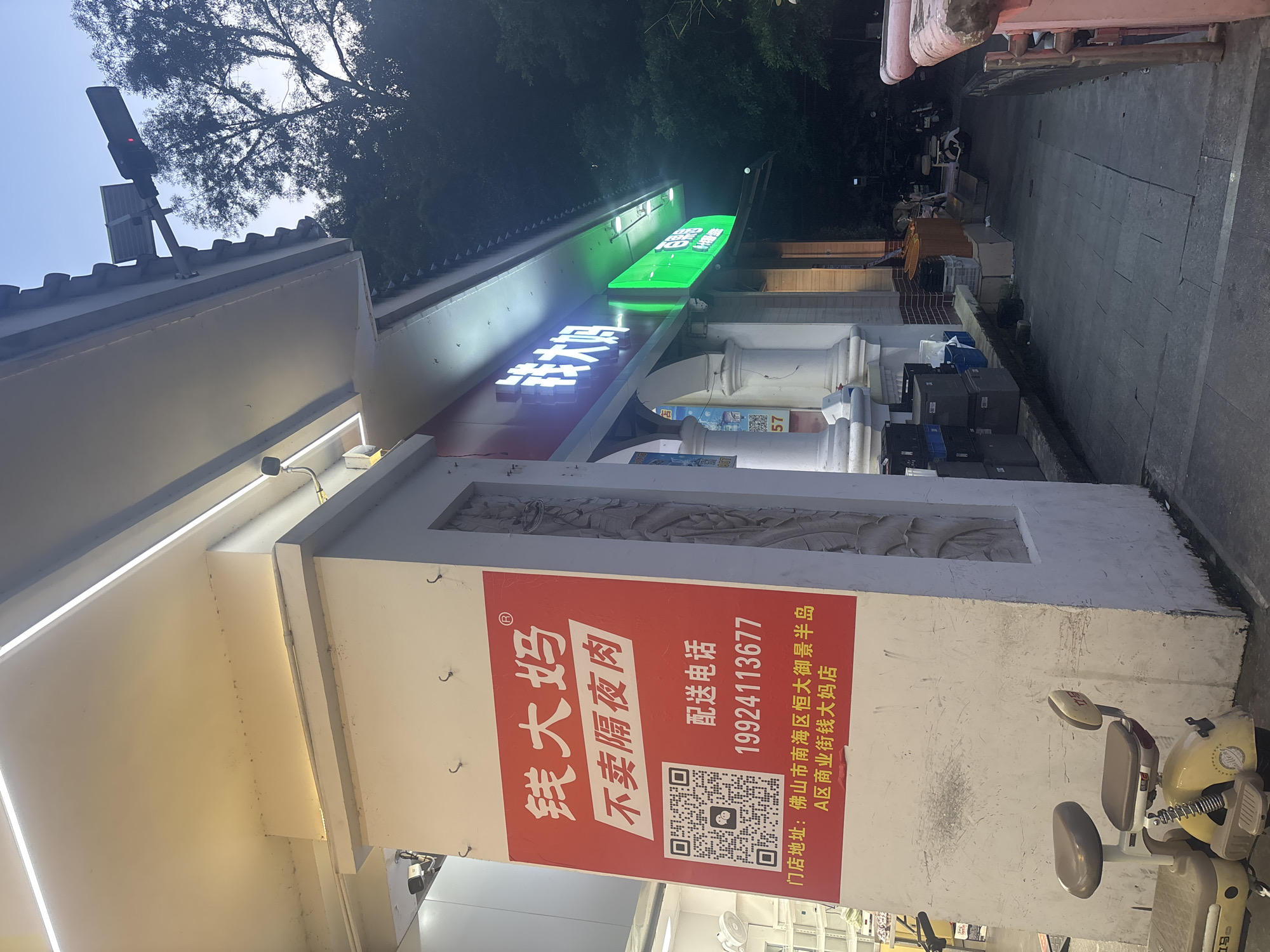 The image shows the exterior of a store named "钱大妈" (Qian Da Ma) in Nanhai District, China. The store appears to be a grocery or convenience store, specializing in fresh produce and possibly meats, as indicated by the text "不卖隔夜肉" (do not sell overnight meat). A large red sign features the store's name, a QR code, a phone number (19924113677) for delivery, and the store's address: "佛山市南海区恒大御景半岛A区商业街钱大妈店" (Qian Da Ma Store, Commercial Street, Zone A, Evergrande Imperial Scenic Peninsula, Nanhai District, Foshan City). The store has a brightly lit facade with the name displayed in illuminated white characters on a dark background above a green illuminated sign. In front of the store, there is a small yellow electric scooter. The surrounding environment suggests an urban setting with trees visible in the background and a paved walkway. The lighting indicates it is likely dusk or early evening.