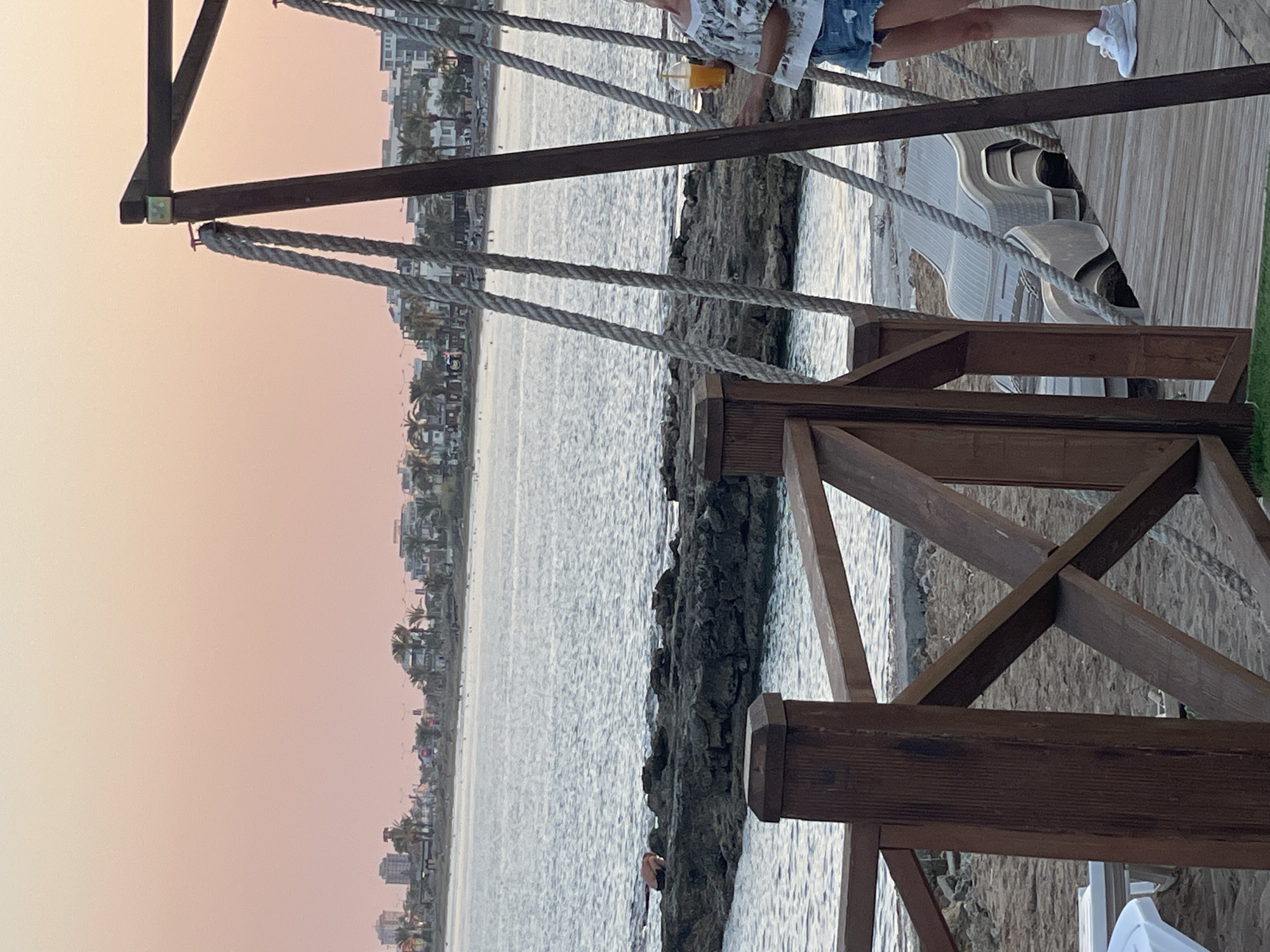 The image depicts a seaside location at dusk, likely in Trikomo, Northern Cyprus. In the foreground, a wooden railing with diagonal supports frames a sandy beach with stacked lounge chairs. To the right, a woman stands on a wooden walkway, holding a drink in a plastic cup and wearing denim shorts and a patterned top. The background features the calm sea with a rocky shoreline and a distant city skyline with buildings and palm trees. The sky is a soft pinkish hue, indicative of sunset.