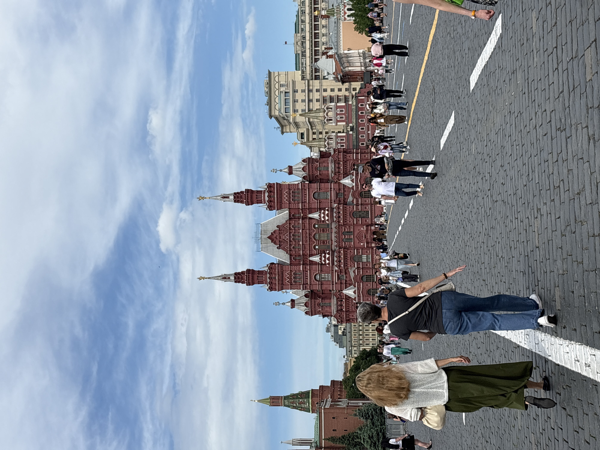 The image captures a scene in Moscow, Russia, specifically in Red Square, featuring the State Historical Museum. The photograph is taken at ground level, with the view skewed at an angle. The sky is partly cloudy. The foreground of the image shows a portion of the paved ground, and numerous people are walking in different directions. Two individuals are more prominent: a woman with blonde hair wearing a light-colored top and a green skirt, and a person in a dark shirt and jeans. The red brick facade of the State Historical Museum is visible and a building complex beyond the museum.