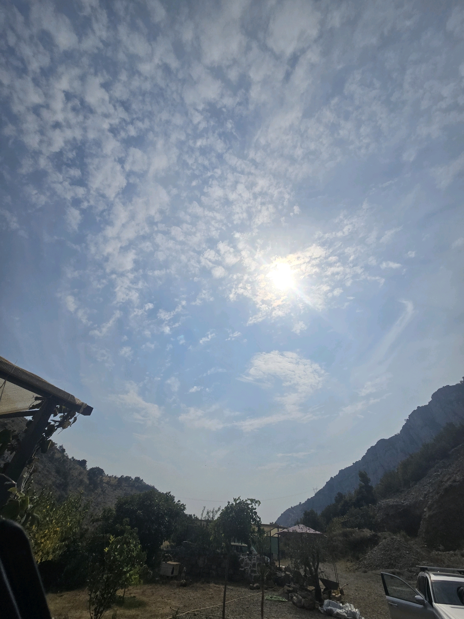 The image depicts a sunny day in a rural or mountainous area, likely on the outskirts of Podgorica, Montenegro, characterized by a clear blue sky dotted with scattered white clouds. The sun is prominent, casting a bright light and creating a lens flare in the upper center of the frame. In the foreground, to the left, a portion of a dark-colored structure with a corrugated roof is visible, suggesting some form of shelter or building. The landscape is dry and arid, with rolling hills and rocky slopes dominating the background. Sparse vegetation, including trees and bushes, can be seen on the hillsides and in the midground. A silver car with its driver's side door open is parked on the right side of the frame. Various objects, including what appears to be a wheelbarrow and some stored materials, are scattered around the ground in the midground, suggesting an ongoing activity or storage area. The overall scene conveys a sense of natural beauty and a somewhat undeveloped environment.