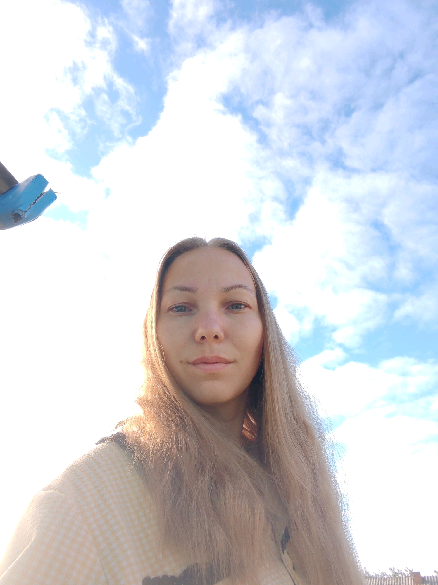 In the city of Chuchlem, Russia, a young woman with long, blonde hair takes a selfie outdoors. She is positioned centrally in the frame, with the majority of the image occupied by a bright, partly cloudy sky. The sky is a vibrant blue with scattered white clouds, suggesting it is daytime and likely sunny. To the upper left of the frame, a portion of a blue object, possibly a clamp or a piece of equipment, is visible against the sky. The woman is wearing a light-colored, possibly yellow, checkered garment. The lower right edge of the image shows a sliver of a wooden fence, indicating an outdoor setting possibly in a rural or suburban area. The lighting is bright, with the sun appearing to be behind the photographer, causing some overexposure at the top of the frame and casting a warm glow on the woman's hair and face.