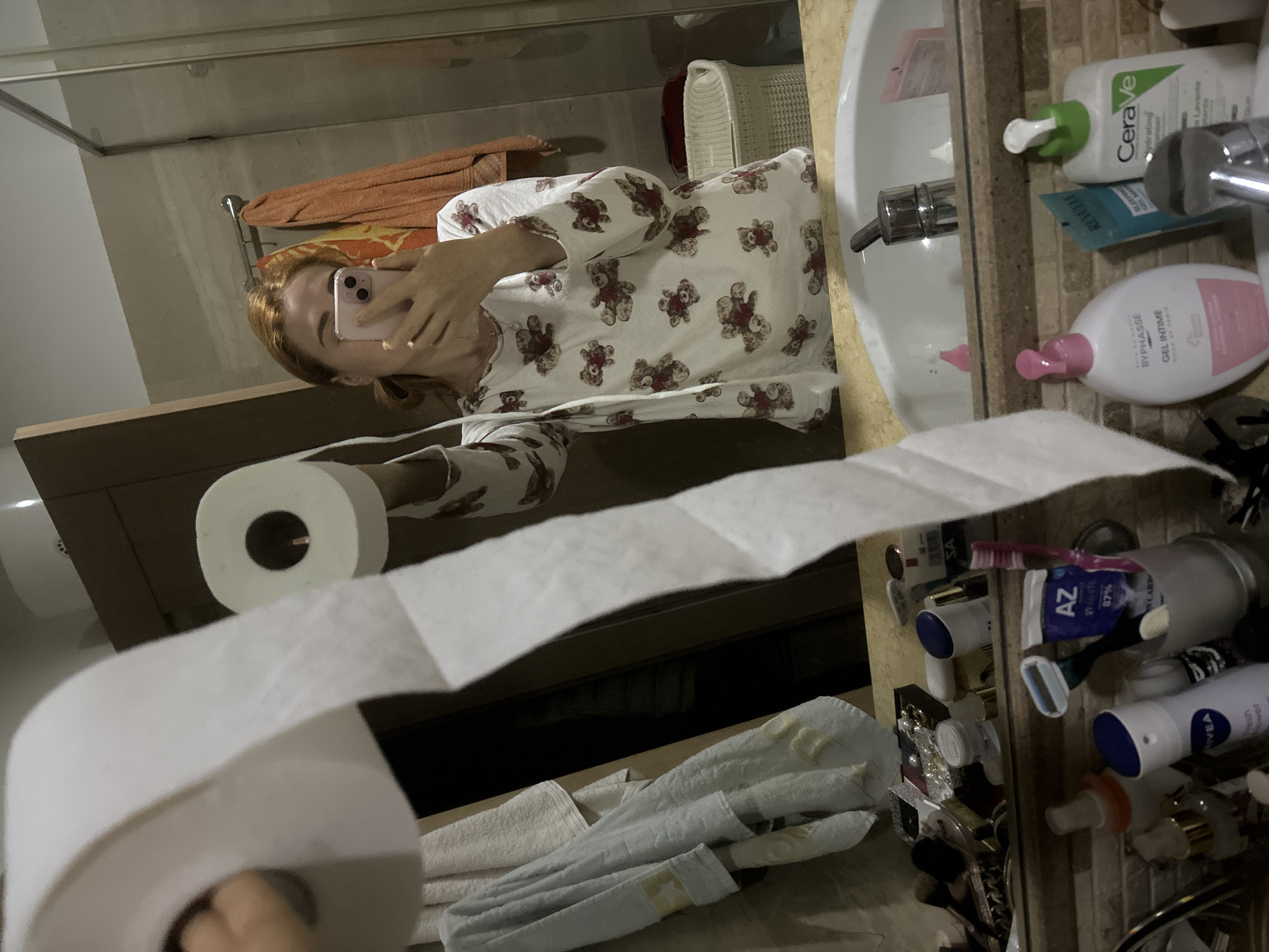 A woman in pajamas is holding a roll of toilet paper in a bathroom and taking a selfie in the mirror. On the countertop, there are toiletries like Nivea deodorant, toothpaste, a razor, Byphasse intimate gel, Revuele sleeping gel mask, and Cerave hydrating cleanser. Towels are visible nearby. The location is likely a private bathroom. The city is not discernible from the image. The country Montenegro is only know from the prompt.