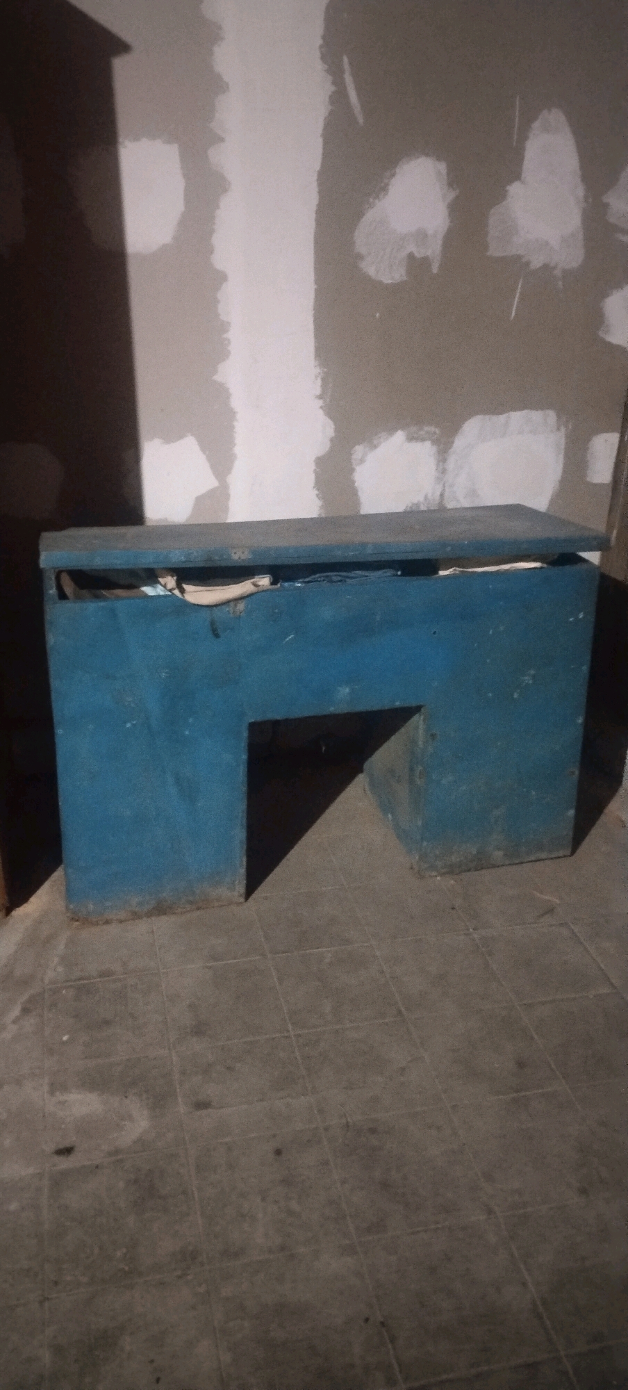 An image shows a blue, cuboid-shaped table with an open top, containing some folded papers and a few dark items. The table is situated in what appears to be an unfinished room with a tiled floor and unpainted walls that exhibit visible patches of plaster. The lighting suggests the presence of artificial illumination, casting shadows on the floor and walls. There are no individuals present in the scene. This scene is likely located in Daleman, Indonesia.