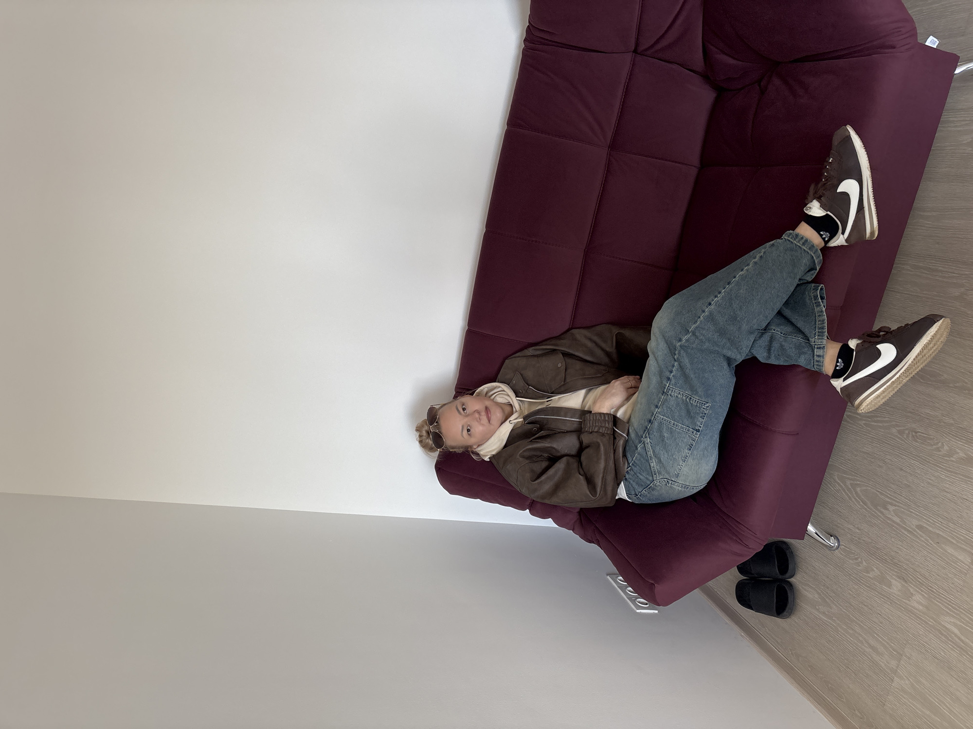 A young adult lies on a plum-colored sofa in a room with a white wall and light wood-tone flooring. The person is wearing blue jeans, a brown jacket, and brown Nike Cortez sneakers with black socks. A pair of black slide sandals are on the floor near the end of the sofa. No information is available in the image regarding the location being in Izhevsk, Russia.