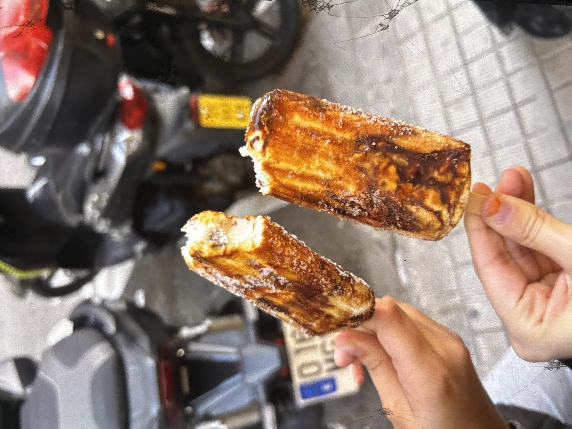 Two churros covered in sugar are held by hands in the foreground. In the background, two motorcycles are parked on a sidewalk. A license plate with the number "016" is visible on one of the motorcycles. The scene appears to be outdoors in a city environment. The churros are golden brown and appear to have been recently fried. The motorcycles are dark in color and appear to be street bikes. The sidewalk is paved with grey tiles. The lighting suggests it is daytime.