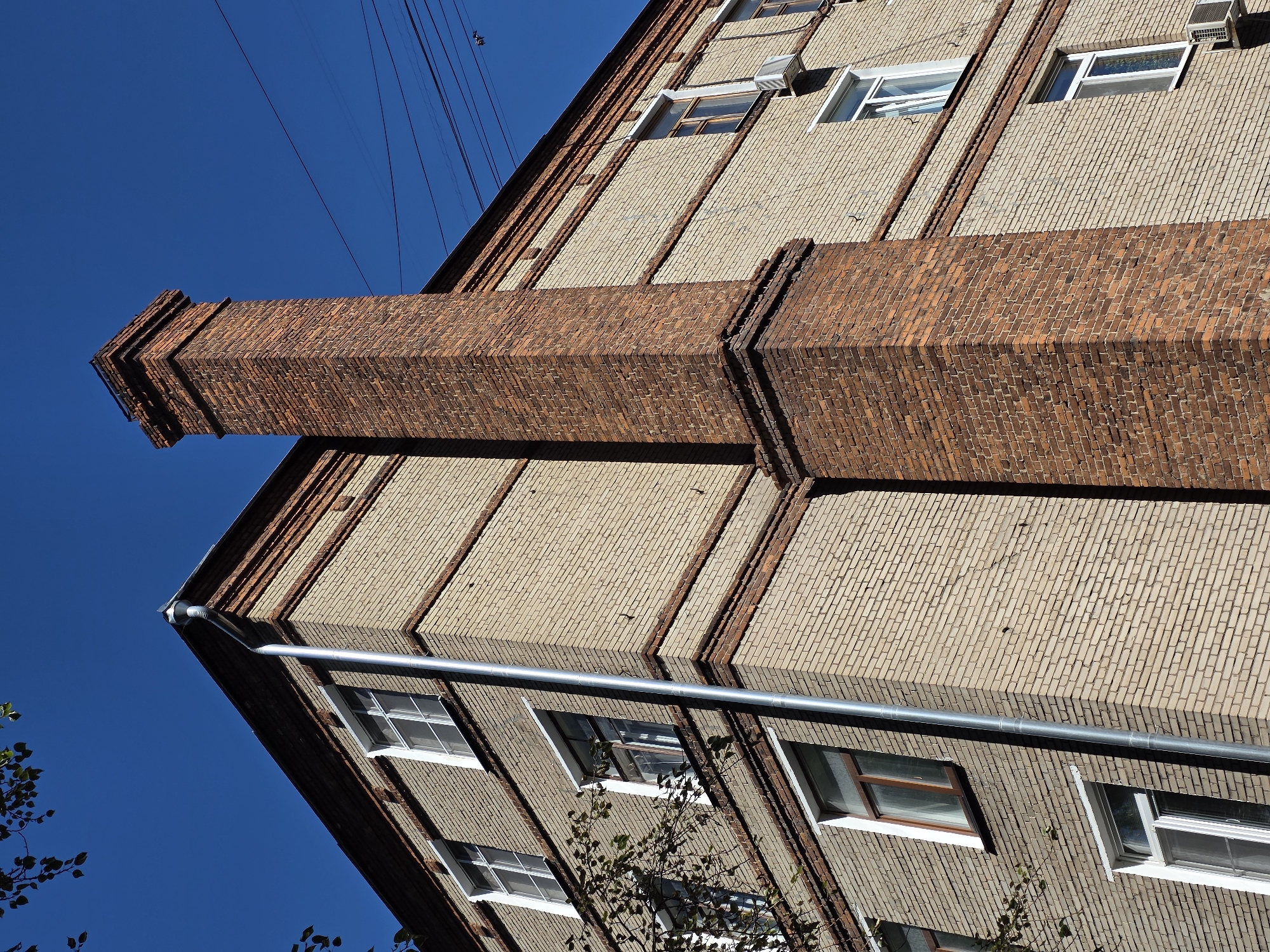 An exterior, low-angle, close-up shot captures the corner of a multi-story building constructed from light-colored bricks with decorative red brick trim. A tall, red brick chimney stands prominently next to the building. Several windows, some with white frames and others with brown frames, are visible on the building's facade. A silver downspout runs vertically along the corner of the building. The sky above is a clear, deep blue. Some tree branches with leaves hang into the frame from the upper left. Electrical wires crisscross the upper right portion of the image against the sky. No people or animals are present in the scene. The overall impression is that of an urban environment on a clear day.