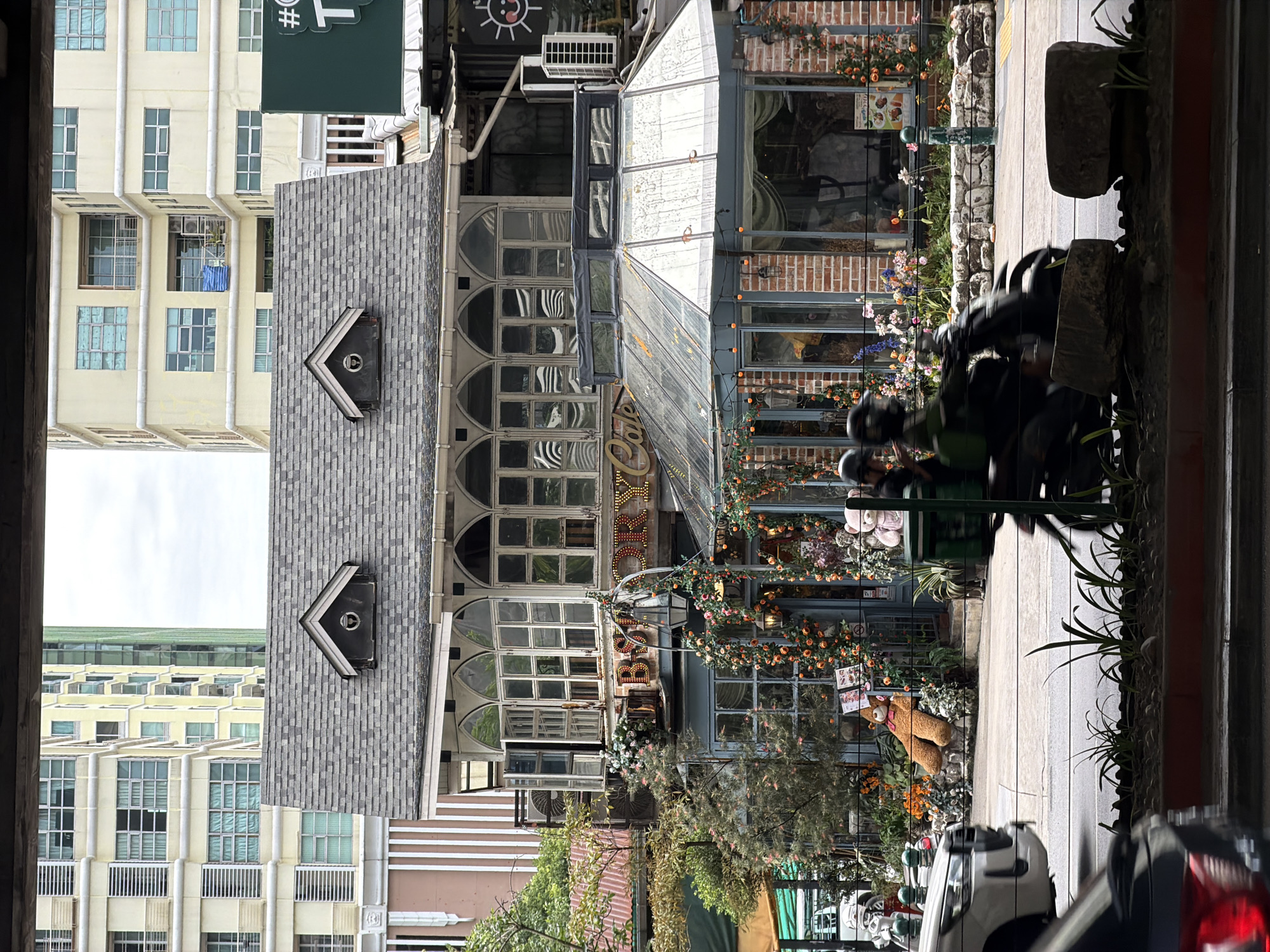 In Bangkok, Thailand, a quaint, European-style cafe with a greenhouse-like extension is situated in front of taller, modern buildings. The cafe features a gray, shingled roof with two dormer windows. Its facade is characterized by large, arched windows with multiple panes, some of which are open. The name "STORY CAFE" is illuminated by warm, yellow lights. The greenhouse section has a glass roof and brick walls.
The cafe is decorated with an abundance of flowers and plants, and two large teddy bears are visible outside, one sitting and one leaning. A stone border with small plants runs along the front of the cafe.
In the foreground, the scene is partially obscured by traffic. A white car is on the left, and to the right, two people on a motorcycle are in motion, their figures blurred by speed. The road is visible, as is a strip of plants in planters. The overall impression is of a charming, European-inspired establishment set against the backdrop of a bustling Asian metropolis.