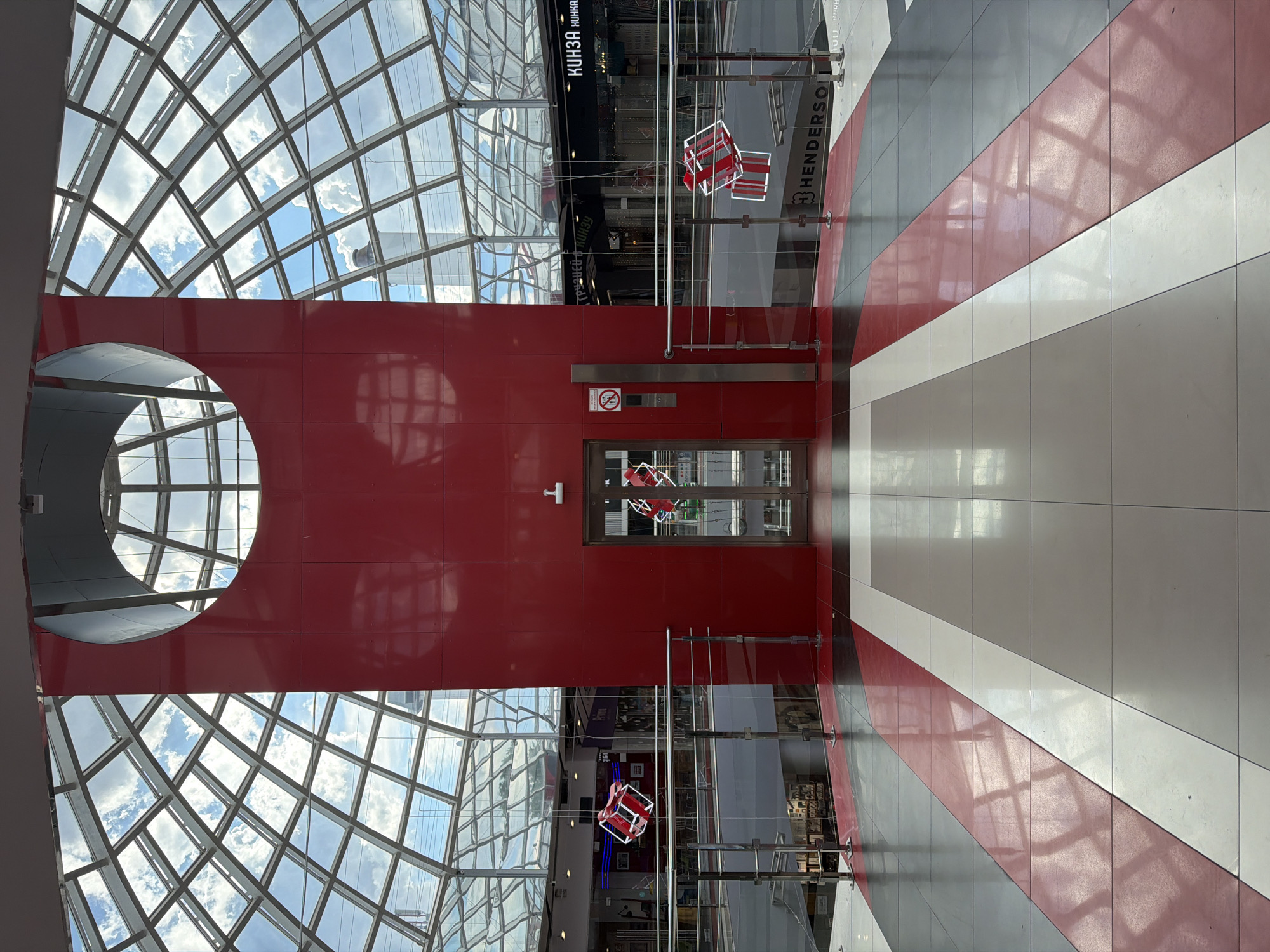 The photograph shows the interior of a building with architectural details. A large red structural element with a circular opening and a rectangular door in the center is the focal point. Above this red structure is a glass grid roof, allowing natural light to enter. Below, the floor has a pattern of alternating gray, white, and red stripes. Handrails run along the edges of the floor pattern. Cubical decorative elements hang from the ceiling. Signage visible in the background indicates the presence of shops or businesses. The building is located in the city of Izhevsk, Russia.