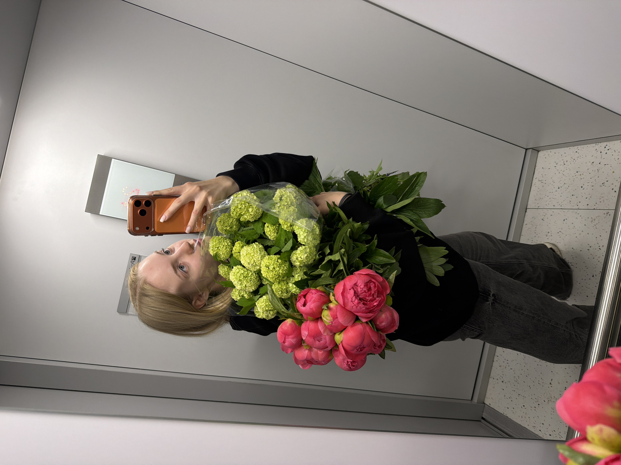 A fair-skinned blonde woman holds a phone in her right hand, taking a selfie in a mirrored elevator. She is wearing a black sweater and gray jeans. She holds a bouquet of green and red peonies in her left arm. The elevator has white walls and a speckled white and gray floor. A small rectangular metal box is attached to the wall above her left shoulder. A metal handrail is visible on the right side of the mirrored surface.