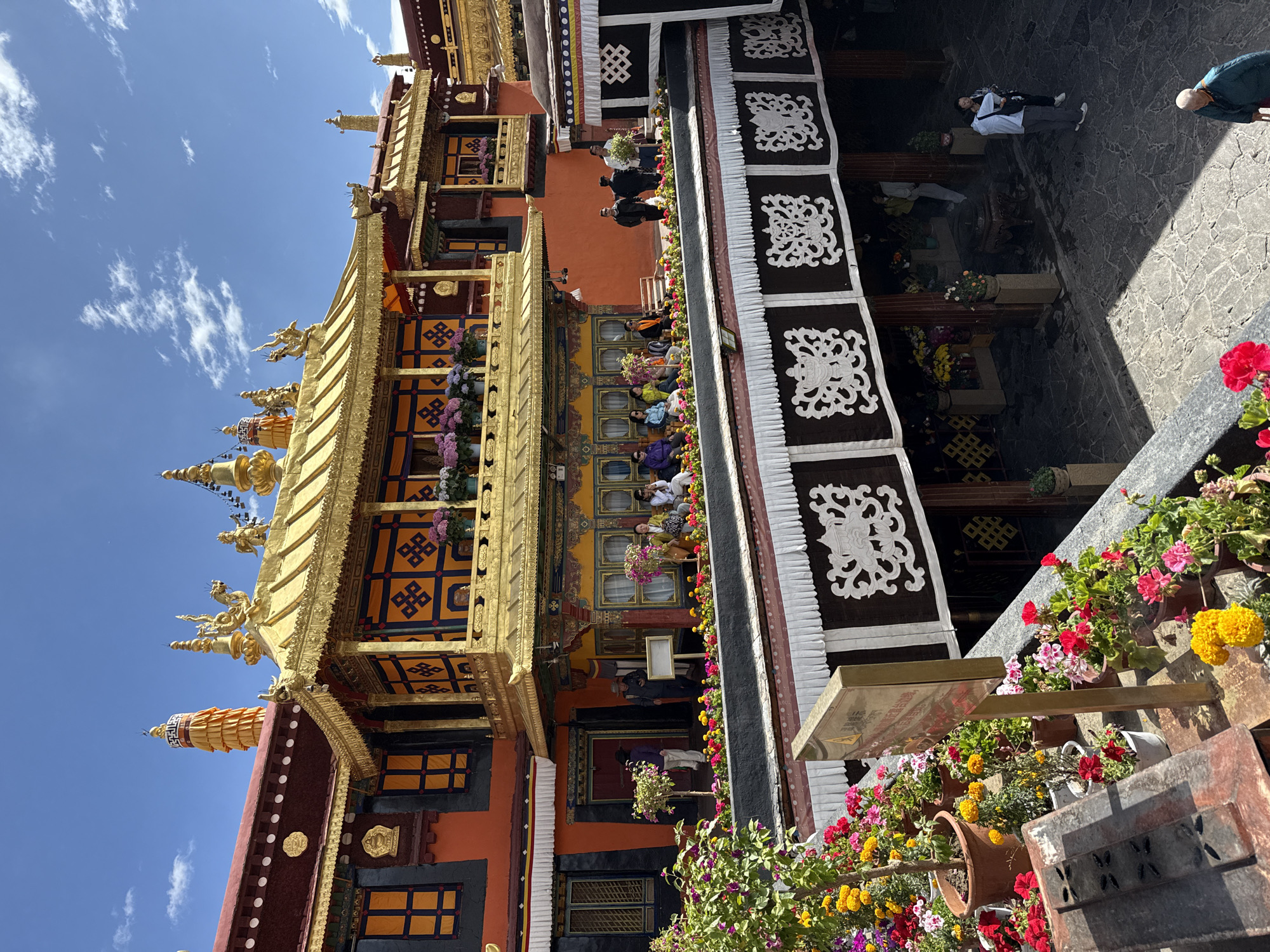 The image depicts a multi-tiered building with a prominent gold roof, observed in Chengguan District, Lhasa, Tibet Autonomous Region, China. The structure features traditional Tibetan architectural elements, including decorative carvings and ornate detailing. People can be observed both inside and outside the building. The foreground shows numerous potted plants with colorful flowers. A blue sky with scattered clouds is visible above the building.