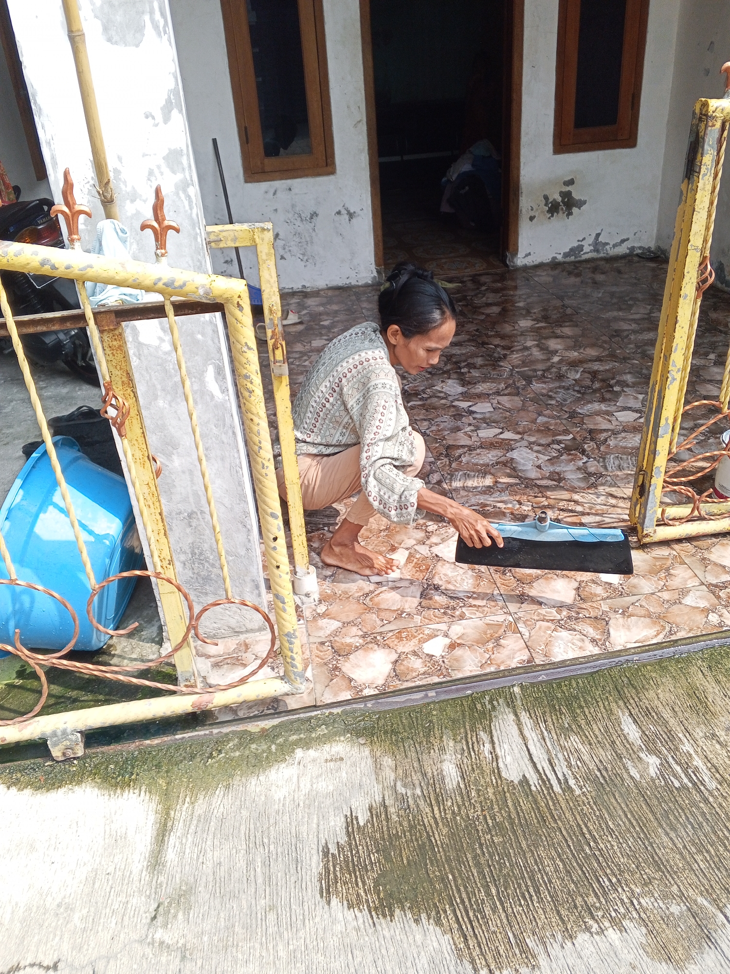 In a courtyard in Ciruas, Indonesia, a woman kneels on a tiled floor and uses a floor squeegee to dry the ground. She is wearing a light-colored, patterned long-sleeved shirt and light brown pants. A large blue plastic tub is positioned to her left, behind a yellow metal fence. The courtyard features patterned floor tiles and a concrete area at the bottom of the frame. A doorway and windows of a building are visible in the background.