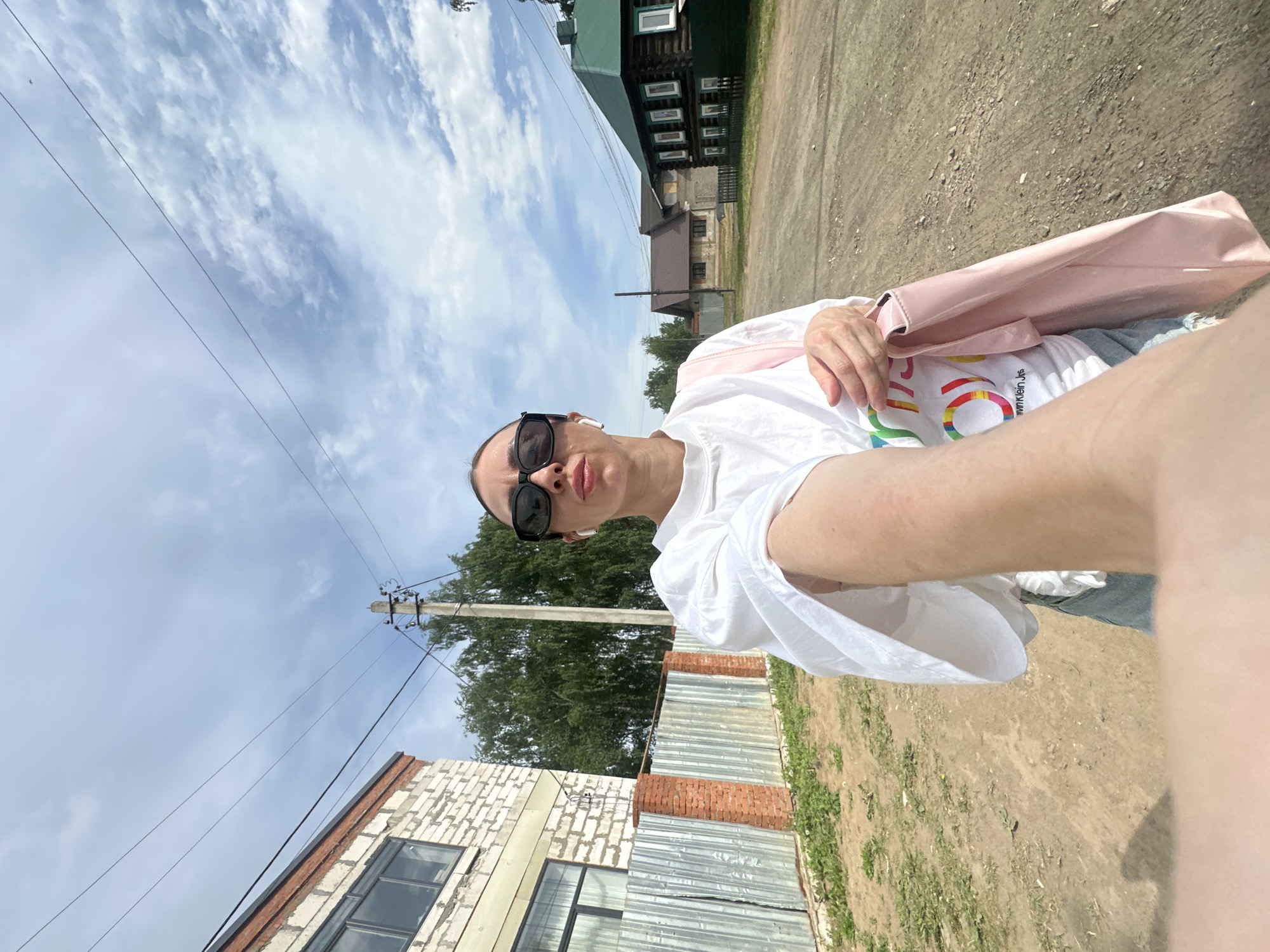 A selfie-style photograph depicts a person wearing sunglasses, a white t-shirt with a rainbow design, and earbuds. The person is holding a pink bag. The image appears to be taken outdoors on a dirt road in what seems to be a rural or suburban area. In the background, there are buildings, including a brick structure and a traditional wooden house with detailed window frames. Utility poles and wires are visible against a cloudy sky. Based on the environment, the location may be Votkinsk, Russia.