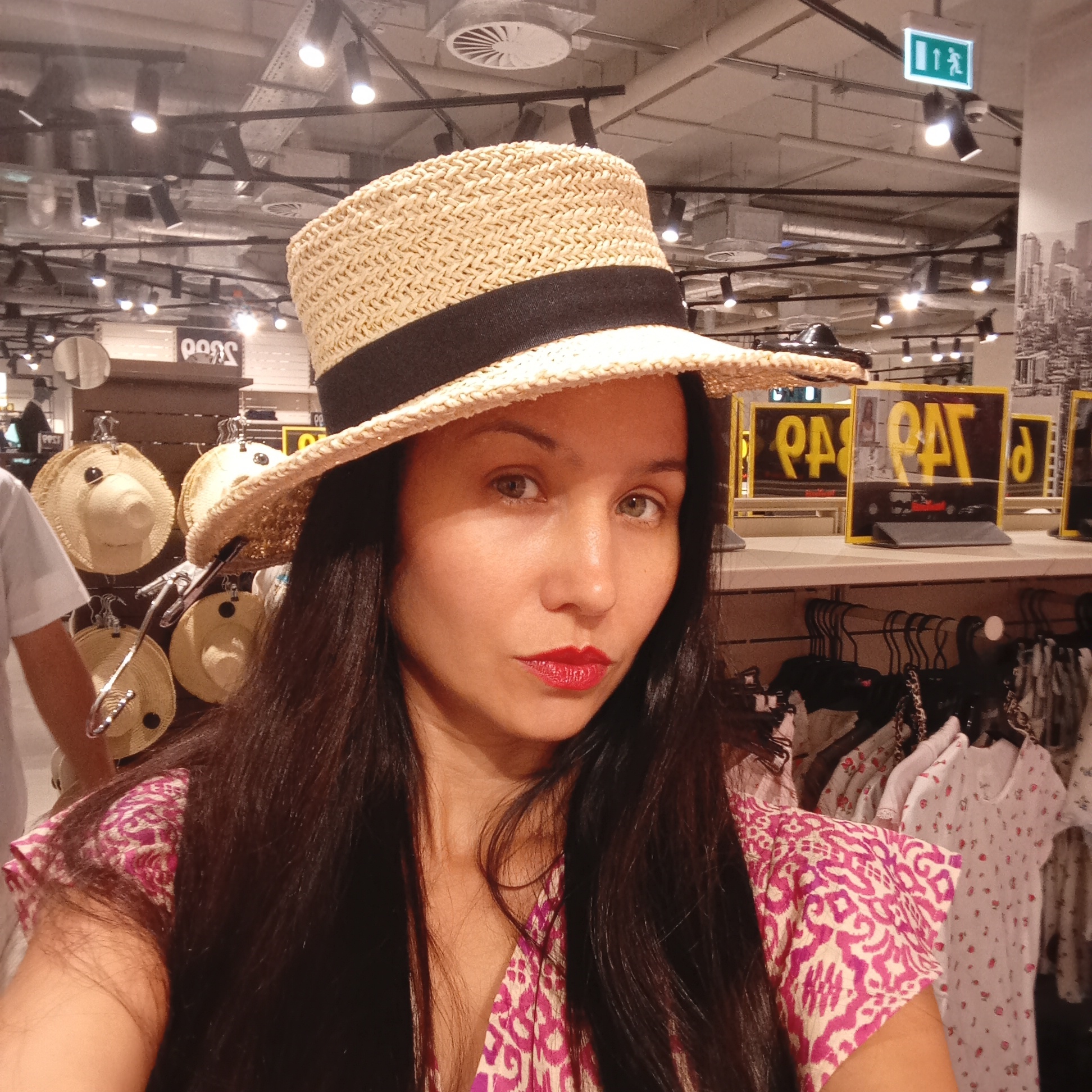 The image shows a close-up, selfie-style shot of a woman wearing a straw hat and a patterned dress. She has dark hair and is wearing red lipstick. The background appears to be an indoor retail environment, possibly a clothing store. Racks of clothing are visible, along with shelves displaying hats and signage. The lighting suggests an artificial, possibly fluorescent, light source. There is no indication that this location is in Sochi, Russia.