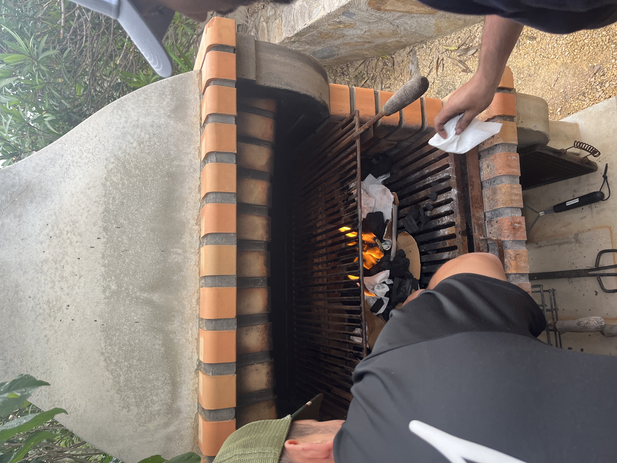 The image depicts an outdoor grilling area, likely in the city of el Poble Nou de Benitatxell / Benitachell, Spain, given the architectural style and vegetation. A tall, brick barbecue grill dominates the frame, containing burning charcoal or wood. Two individuals are visible. One is holding a white cloth and appears to be tending to the grill. The other person is closer to the camera, wearing a black shirt with a white design and a green hat. Various grilling tools and accessories are scattered near the grill. The area seems to be on a patio or terrace with stone or concrete flooring. Greenery and trees are visible in the background.
