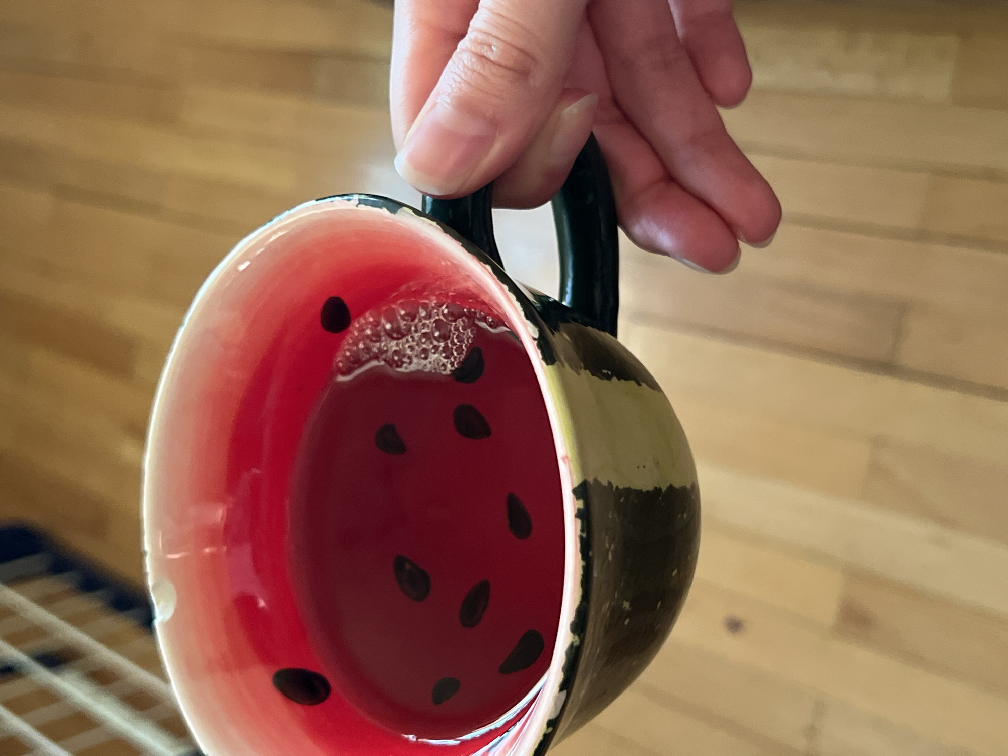 A hand holds a watermelon-themed mug filled with a reddish liquid. The mug is painted red inside with black seed shapes, resembling the inside of a watermelon, while the exterior is striped in green and dark green, mimicking a watermelon rind. There are some bubbles on the liquid's surface. The background is a light-colored, wooden floor. A metal grate is partially visible in the lower left corner. The location of the scene is not discernible from the image.