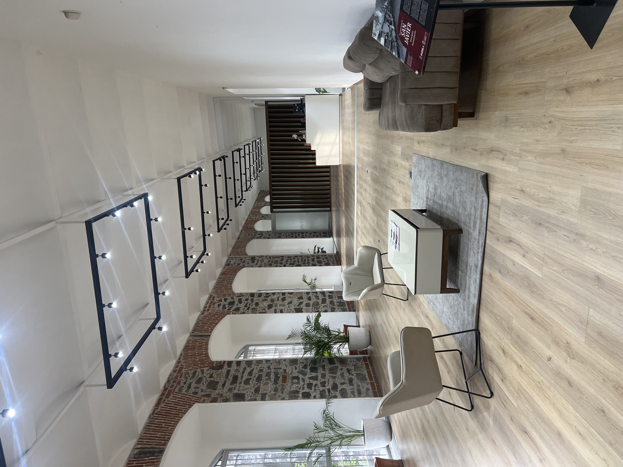 This image captures the interior of a spacious and well-lit building, likely a lobby, reception area, or public exhibition space, with a blend of historical and modern architectural elements. The perspective is from a slightly elevated position, looking down the length of the hall.
The left side of the hall features a long wall with exposed brick and stone arches, suggesting a historic structure. Through the arched openings, windows with grates are visible, allowing natural light to enter. Potted palm plants are placed at the base of some of these arches. The upper section of this wall and the high ceiling are painted white. Modern industrial-style lighting fixtures, consisting of rectangular black frames with multiple exposed bulbs, are suspended from the ceiling, running down the length of the hall. A smoke detector is visible on the white ceiling.
On the right side of the hall, there is a seating area with light wooden flooring. A large, multi-sectioned grey sofa is positioned against the wall towards the far end. Closer to the foreground, a grey rectangular rug defines a space with two cream-colored armchairs with slender black metal frames, facing a rectangular white coffee table.
In the background, a modern wooden staircase with dark treads ascends, leading to an upper level. Two individuals are partially visible on the staircase, one appearing to ascend and another person's head visible further up.
A tall display stand with an information sign is located next to the sofa. Visible text on the sign reads "SAN JAVIER" in large letters, followed by "PROTECCIÓN PATRIMONIAL" and "PUEBLA," indicating the location is Puebla, Mexico, and possibly pertains to heritage protection or a historical site named San Javier. The overall ambiance suggests daytime, given the bright interior illumination from both natural light and artificial fixtures. The space appears clean, organized, and quietly inviting.