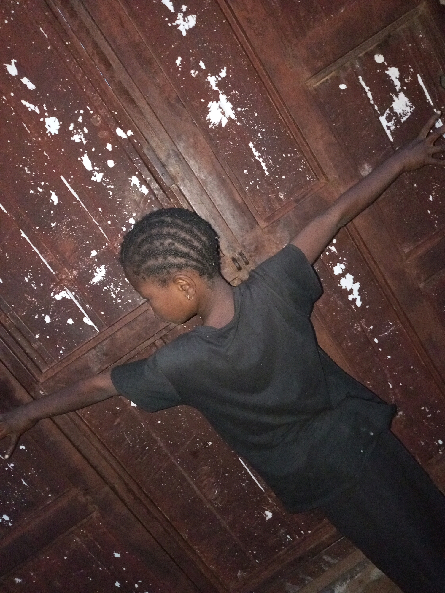 A young person with braided hair and wearing a black t-shirt and black pants is standing with arms outstretched against a dark brown wooden surface. The wooden surface appears to be a door or wall, with visible seams and an uneven, weathered texture. White specks, possibly paint splatters, are scattered across the wooden surface. The person's head is tilted down and to the side, obscuring their face. The lighting is dim, casting shadows and creating a slightly surreal atmosphere. The image appears to be taken indoors, possibly in a dimly lit room or an outdoor area with limited light.