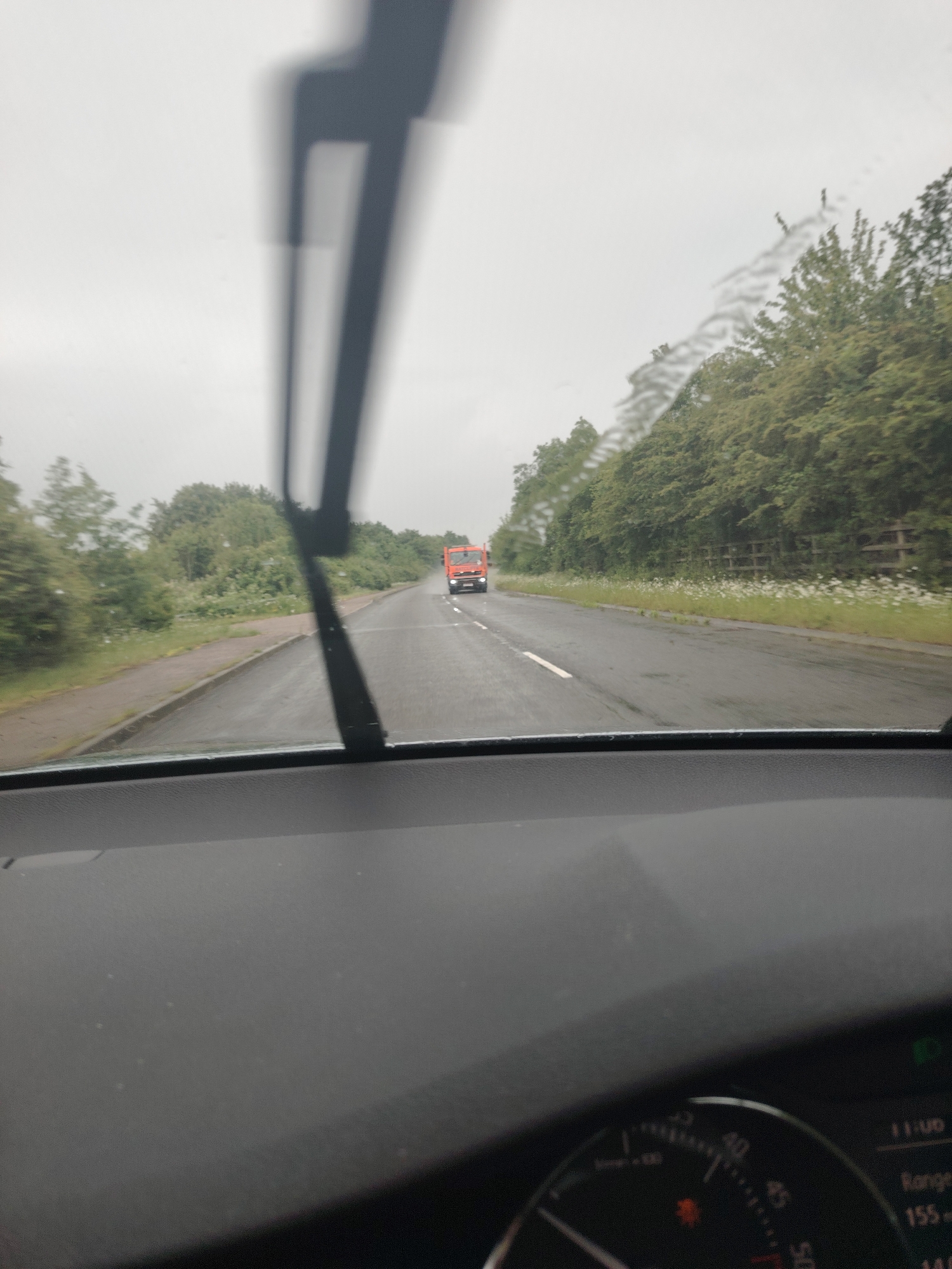 The image captures a road scene from the interior perspective of a moving vehicle, likely in Huntingdonshire, United Kingdom. The foreground shows the car's dashboard and the lower part of the windshield, with prominent dark windshield wipers blurred across the view, indicating motion or a recent swipe. Water streaks are visible on the windshield, suggesting rain or recent wet weather, which is consistent with the damp appearance of the road.
An overcast, bright grey sky dominates the upper portion of the frame. On both sides of the two-lane road, lush green trees and dense hedges line the route, with a paved sidewalk visible on the left. In the distance, a distinct bright red truck, possibly a utility or refuse vehicle, is approaching on the opposite side of the road. White road markings are visible on the asphalt.
Inside the car, a portion of the dashboard and instrument cluster is visible in the bottom right. A digital display shows the time "11:00" and a "Range 155" (likely miles or kilometers remaining). Part of a speedometer or tachometer is visible, indicating speed markers like "20," "30," "40," and "50," along with an illuminated amber warning light. The overall impression is a somewhat dreary, wet drive during late morning hours.