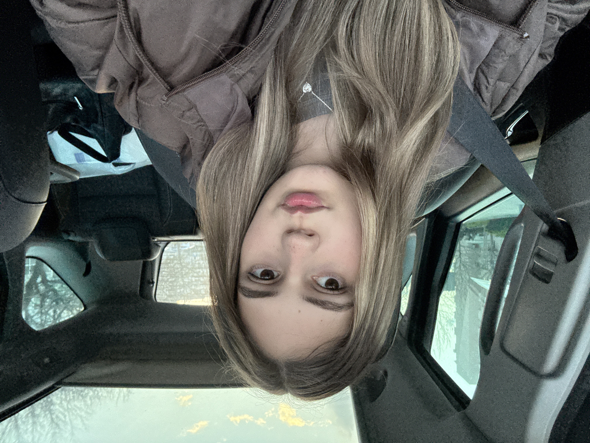 The image is a selfie taken from inside a car. A young person with light brown hair is the central subject. They are wearing a brown jacket and a seatbelt. A silver necklace with a small pendant is visible. The interior of the car is visible, including the dashboard and the windows. Through the windows, the exterior environment can be seen, including what appears to be buildings and trees. There is no indication to confirm that the location is Panagyurishte, Bulgaria.