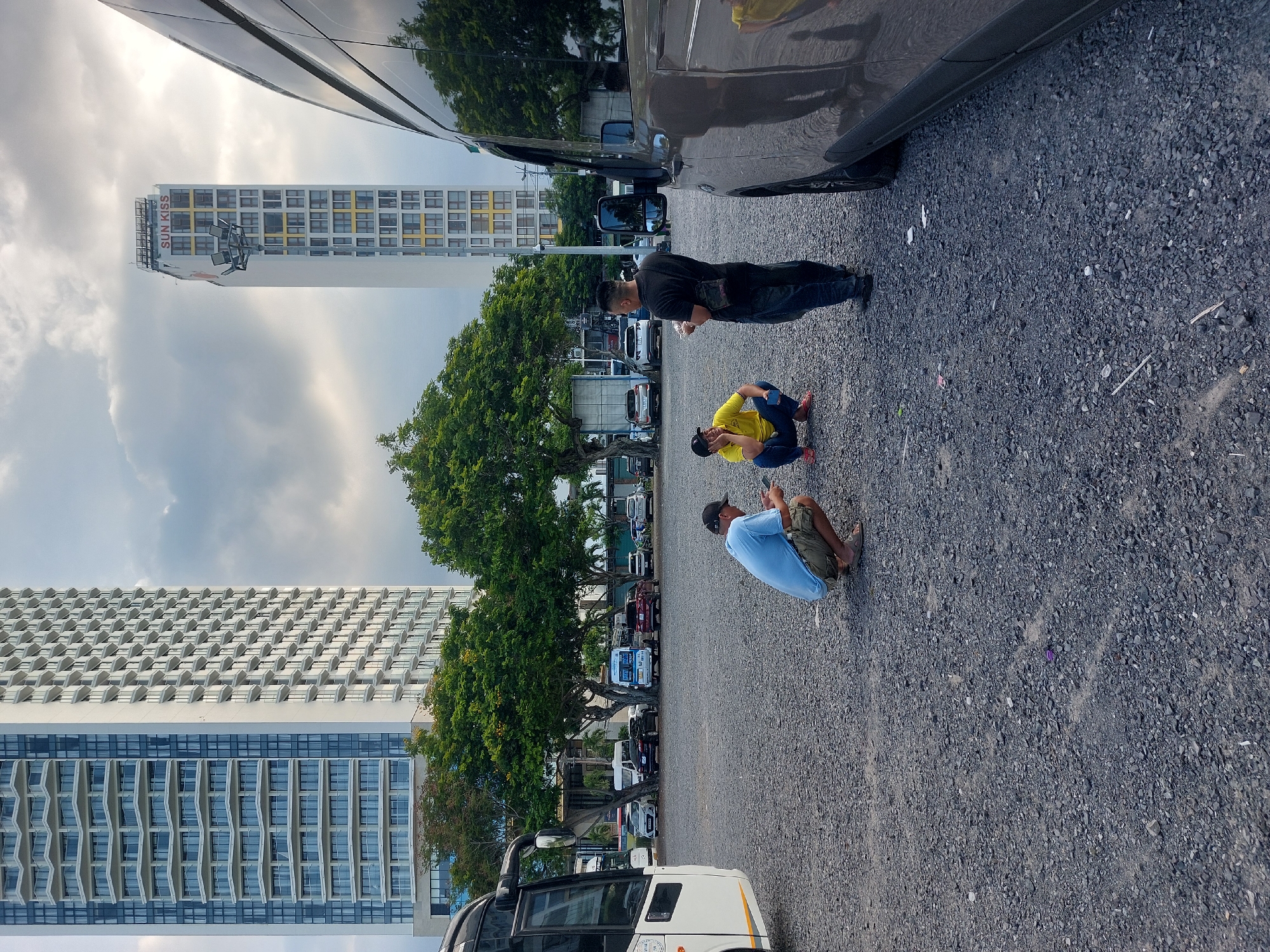 Three men are gathered in a gravel parking lot, with two tall buildings in the background, one with a "SUN KISS" sign at the top. The building on the left is a white high-rise with many balconies, and the building on the right is a narrower, white high-rise with yellow accents. There are trees and several parked vehicles, including vans and cars, behind the men. A large, dark-colored van is parked in the foreground to the right.
The man on the left is wearing a light blue t-shirt and khaki shorts, crouching down and looking at his phone. In the center, a man in a bright yellow t-shirt and blue pants is also crouching, looking towards the man on the left. To the right, a man in a black t-shirt and dark pants stands facing the other two, holding a small white object in his hand. The ground is covered in dark gray gravel and small rocks. The sky is overcast with clouds. The scene appears to be in a commercial or parking area in Nha Trang Ward, Vietnam, given the architectural style of the buildings and the presence of many parked vehicles.