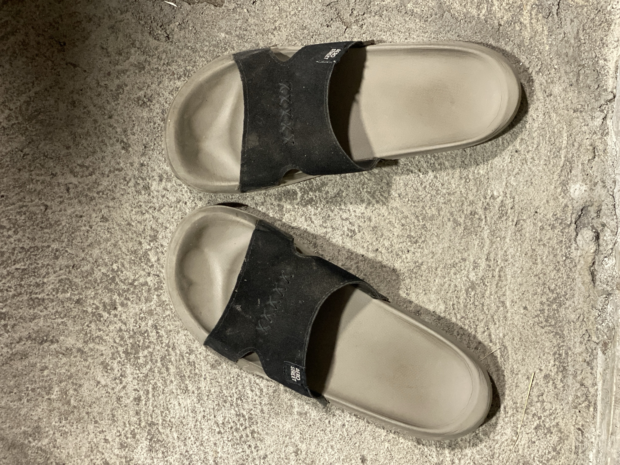 A pair of worn, tan-colored slide sandals with black straps are positioned on a textured concrete surface. The straps have a decorative "XXXX" stitching pattern and a small white label reading "AERO STREET" on the left sandal. The concrete appears to be outdoors, possibly a sidewalk or pathway, with some debris and shadows suggesting it may be evening or a shaded area. The scene is located in Kasihan, Indonesia.