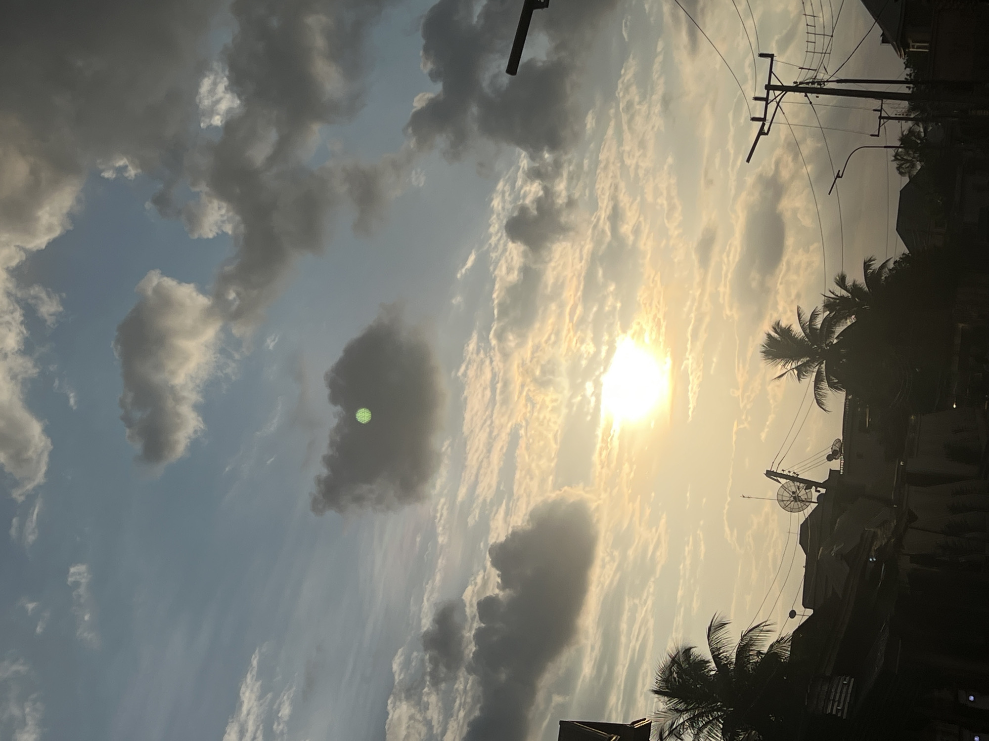 The image captures a dramatic sky during what appears to be sunset or sunrise, with the sun's bright rays piercing through a sky filled with a mix of dark, ominous clouds and lighter, wispy formations. The overall atmosphere is one of transition and perhaps a touch of foreboding due to the heavy cloud cover.
The scene is set in Nkpor, Nigeria. Silhouetted against the vibrant sky are elements of urban or semi-urban life. A prominent feature is a utility pole with a complex network of wires and what seems to be a satellite dish mounted on a building, suggesting a developed area with access to telecommunications. Palm trees are visible, their fronds dark against the luminous sky, indicating a tropical or subtropical climate. The presence of buildings, also in silhouette, further grounds the scene in a populated area.
There is no discernible activity or event taking place; the image focuses solely on the atmospheric conditions and the elements within the landscape. The time of day is clearly indicated by the low angle and intensity of the sun, casting long shadows and creating a warm, golden glow interspersed with cooler blue tones in the sky. The weather is characterized by the dramatic cloud formations, hinting at either the end of a stormy day or the approach of rain, or possibly just the natural spectacle of a colorful sunset.
A notable detail is a small, bright green circular object or light anomaly within one of the dark clouds on the left side of the image. Its origin is unclear and it stands out as an unusual element in the otherwise natural sky. No visible text is present in the image. The emotions evoked are a mix of awe at the natural beauty of the sky and a sense of the everyday presence of infrastructure within a vibrant natural environment.