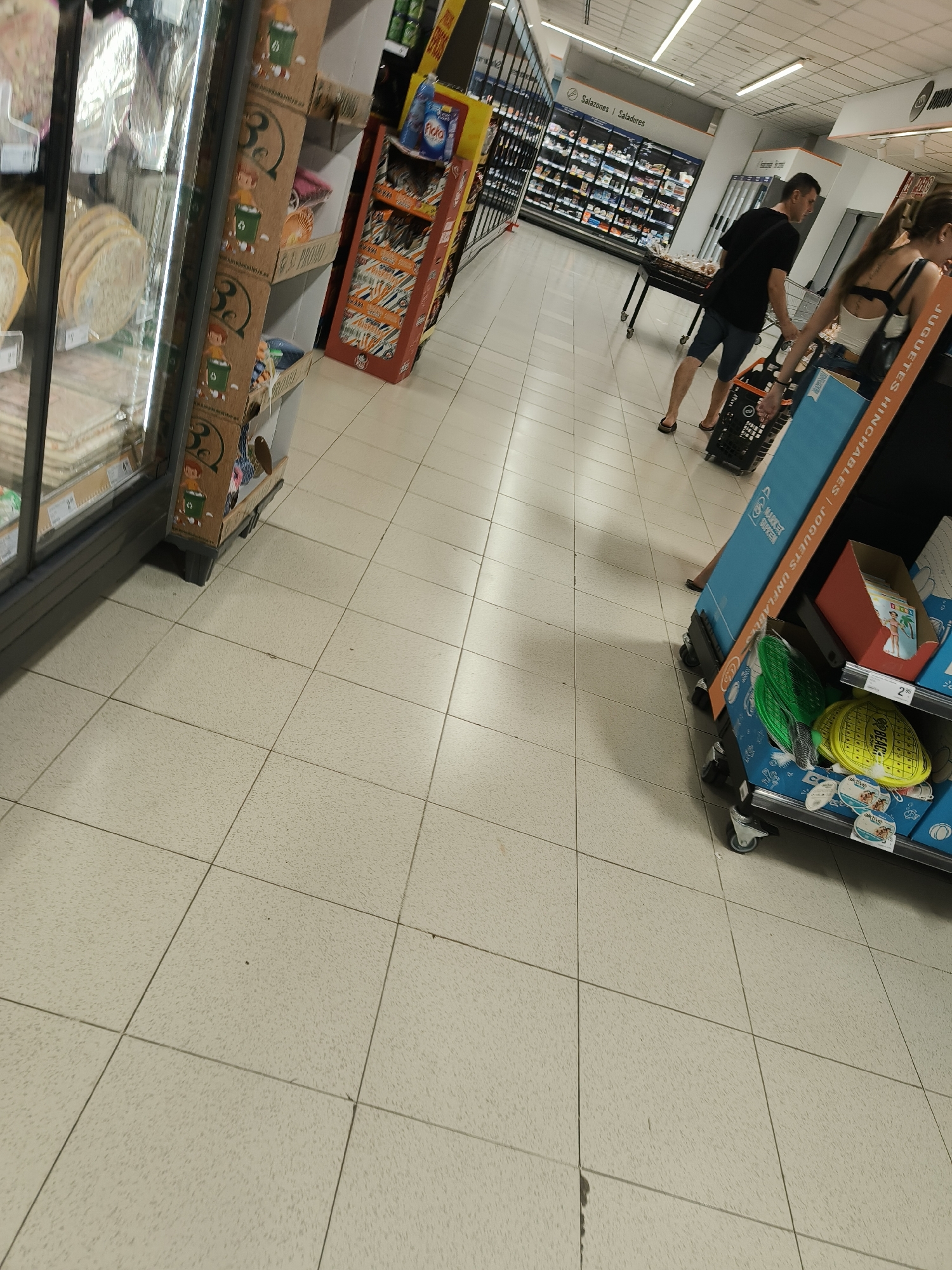 The image shows an interior view of a supermarket in Benimàmet-beniferri, Spain. The scene is dominated by light-colored tiled flooring and displays of various products on shelves and gondolas. In the foreground, to the right, is a display of inflatable toys, including paddleballs and beach balls, some of which are packaged in blue and yellow. Adjacent to this is a red cardboard display containing what appear to be magazines or brochures. In the midground, a man wearing a black t-shirt and shorts is pushing a shopping cart. To his right, a woman with dark hair, wearing a white top and dark straps, is walking away from the camera, also appearing to be shopping. Further in the background, a long row of refrigerated display cases is visible, stocked with various food items. Above the refrigerated cases, a sign reads "Salazones | Saladures". The overall environment suggests a typical supermarket shopping experience.