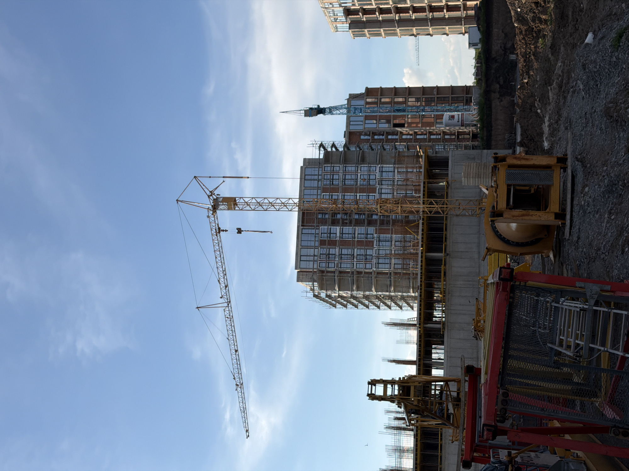 The image shows a construction site in Burgas, Bulgaria. Several high-rise buildings are under construction, with cranes towering above the structures. One crane is yellow, and another, further in the background, is blue. The buildings have scaffolding around them, and construction materials are visible on the ground. The sky is blue with scattered clouds. The foreground includes construction equipment, forms, and rebar. The ground consists of dirt and gravel.