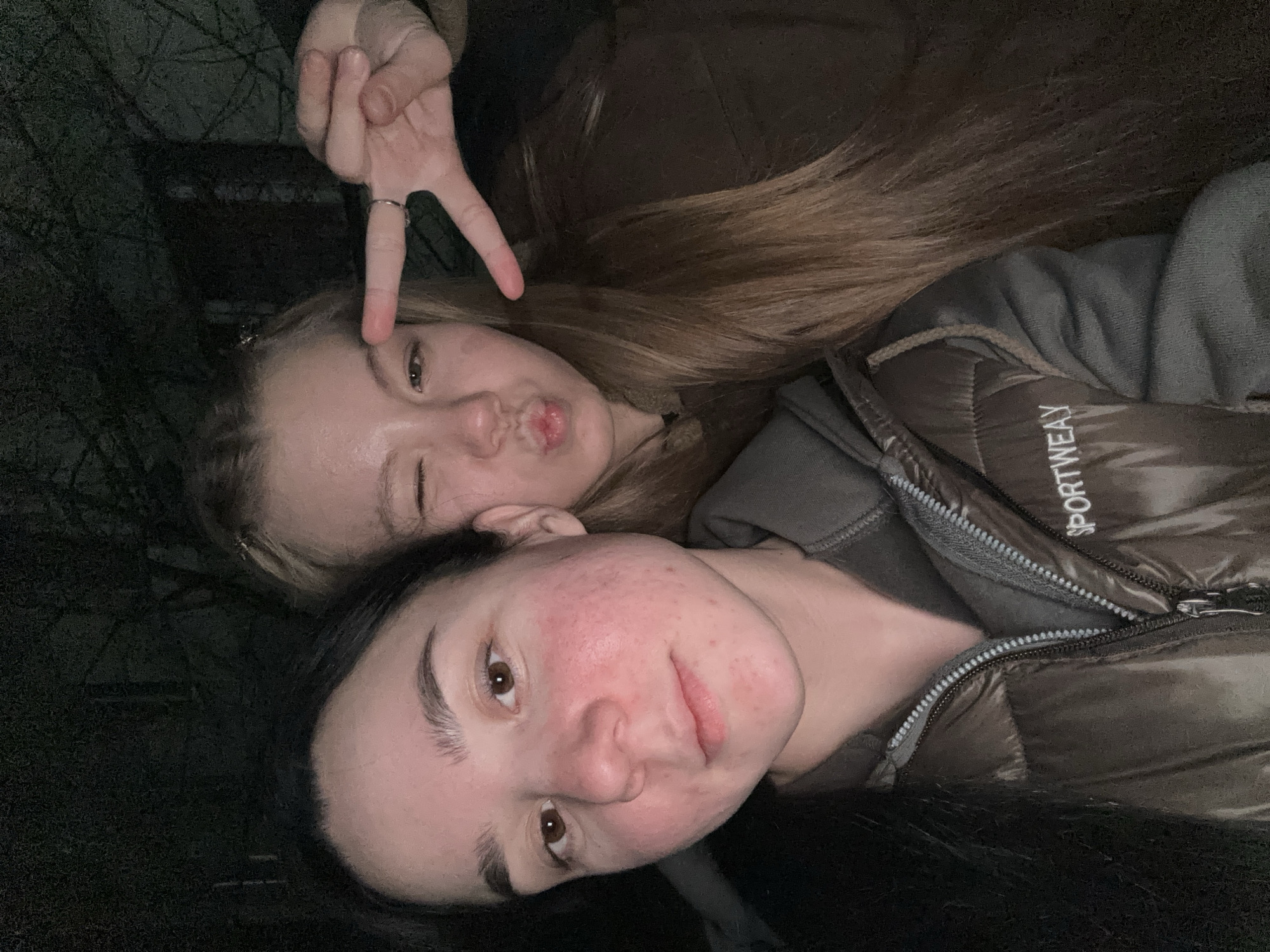 Two young women are posing for a selfie outdoors. The woman in the foreground has dark hair and reddish skin. She is wearing a dark puffer jacket with the word "SPORTWEAR" visible on it. The woman in the background has blonde hair and is making a "duck face" while holding up a peace sign. The lighting is dim, suggesting it might be evening or night. Bare tree branches are visible in the background. The location is potentially the city of Chaikovsky, Russia.