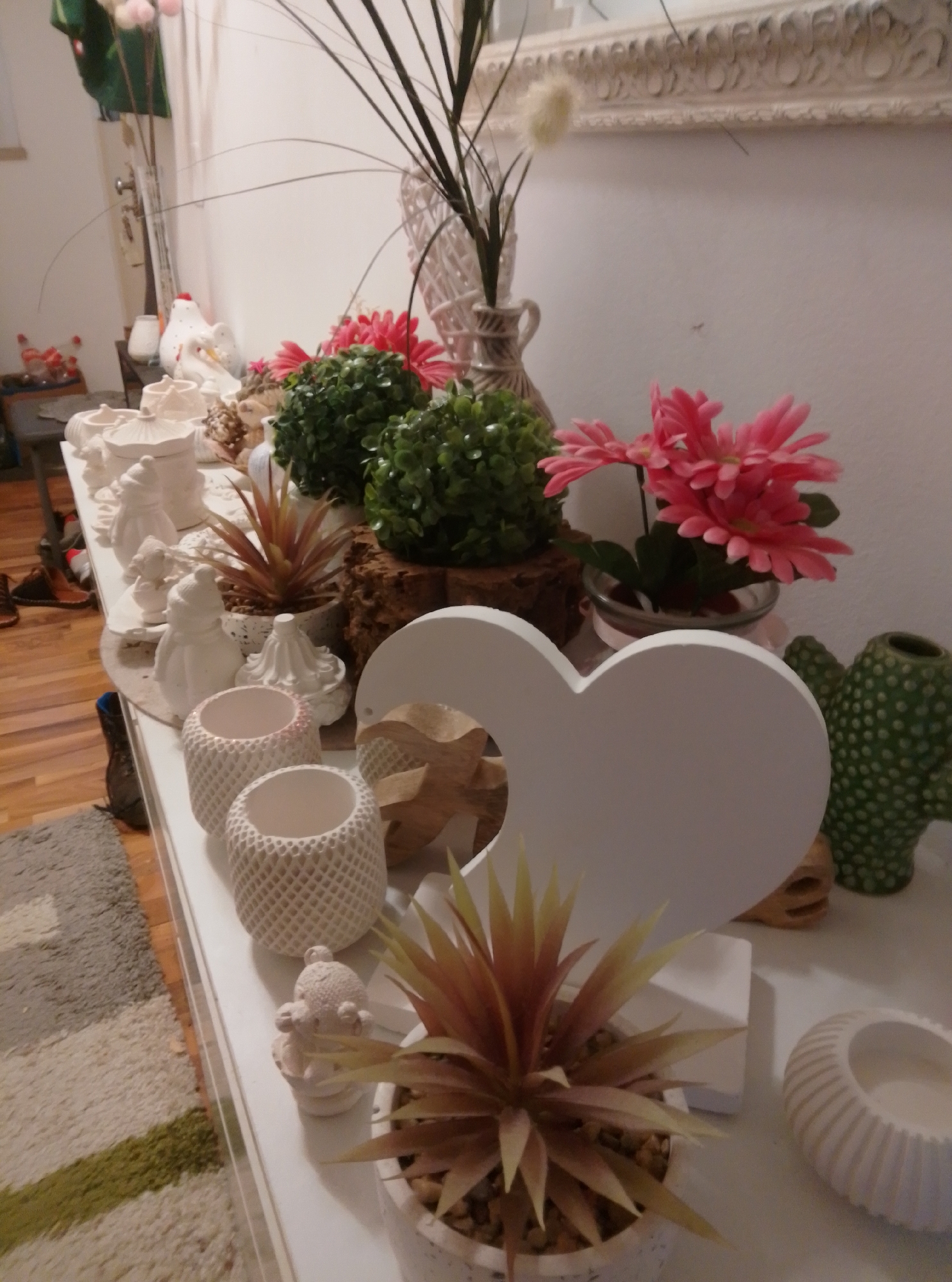 The image depicts a white shelf displaying various decorative items. In the background, a large ornate mirror hangs on a white wall. The shelf is filled with a collection of ceramic figurines, artificial plants, and vases. Prominently featured are several white ceramic figurines, some resembling chickens, others abstract shapes. There are also two green, textured artificial plants and a vase with pink artificial flowers. In the foreground, a white heart-shaped object is visible, along with a planter containing an artificial succulent and two white decorative containers with intricate patterns. A green cactus-shaped ceramic pot sits to the right of the heart. The overall scene suggests an indoor decorative display. The provided image does not contain information about the city of Wedel, Germany, or any people or specific actions.