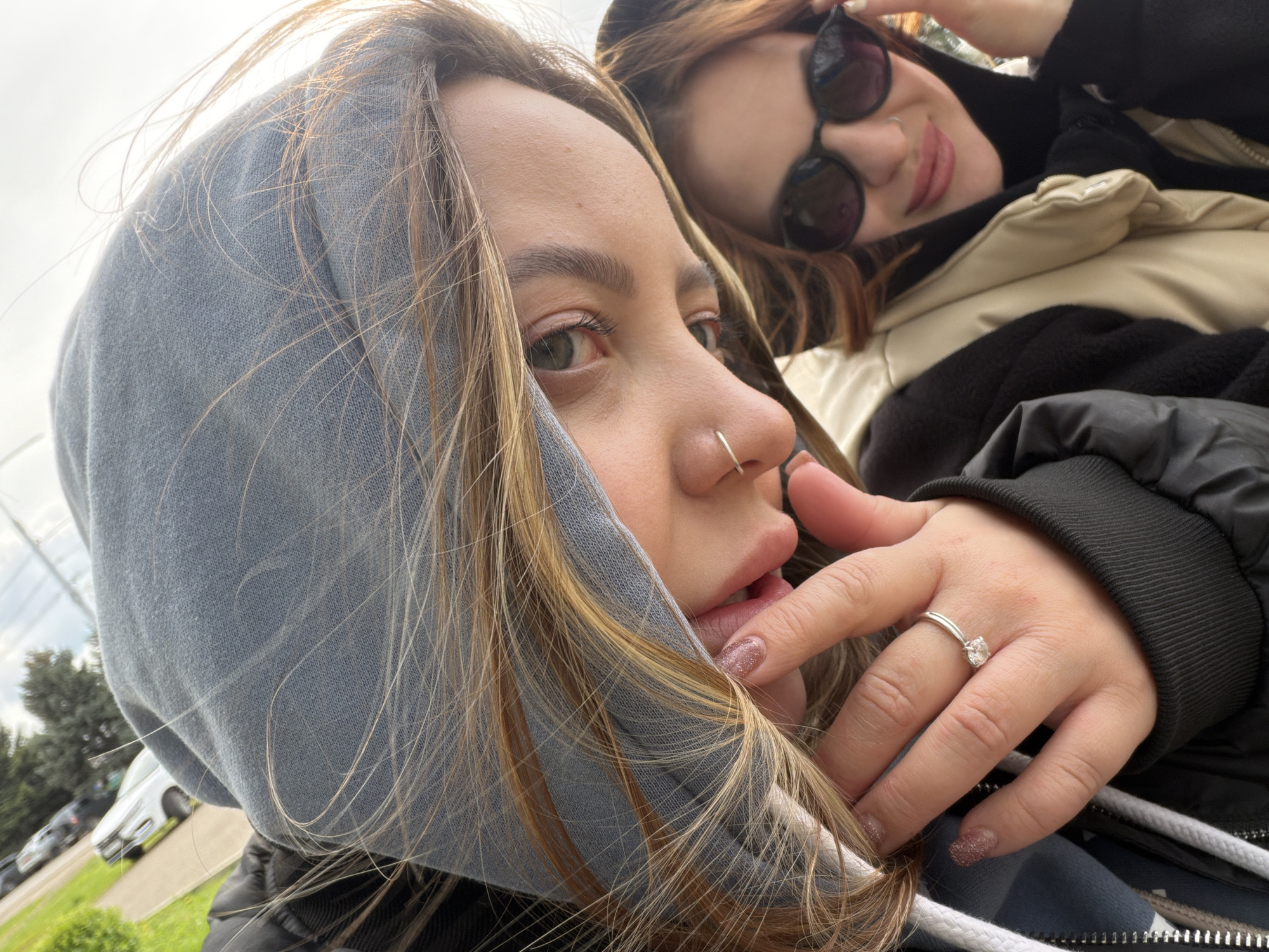 Two young women are posing for a selfie. The woman in the foreground is wearing a gray hoodie with the hood up, a nose ring, and a ring on her finger. She is touching her mouth with her hand. The woman in the background is wearing sunglasses and a black jacket with a beige puffer vest. The background is a blurry mix of trees, a light sky, and parked cars. It is not possible to determine if this image was taken in Novopokrovskaya, Russia.