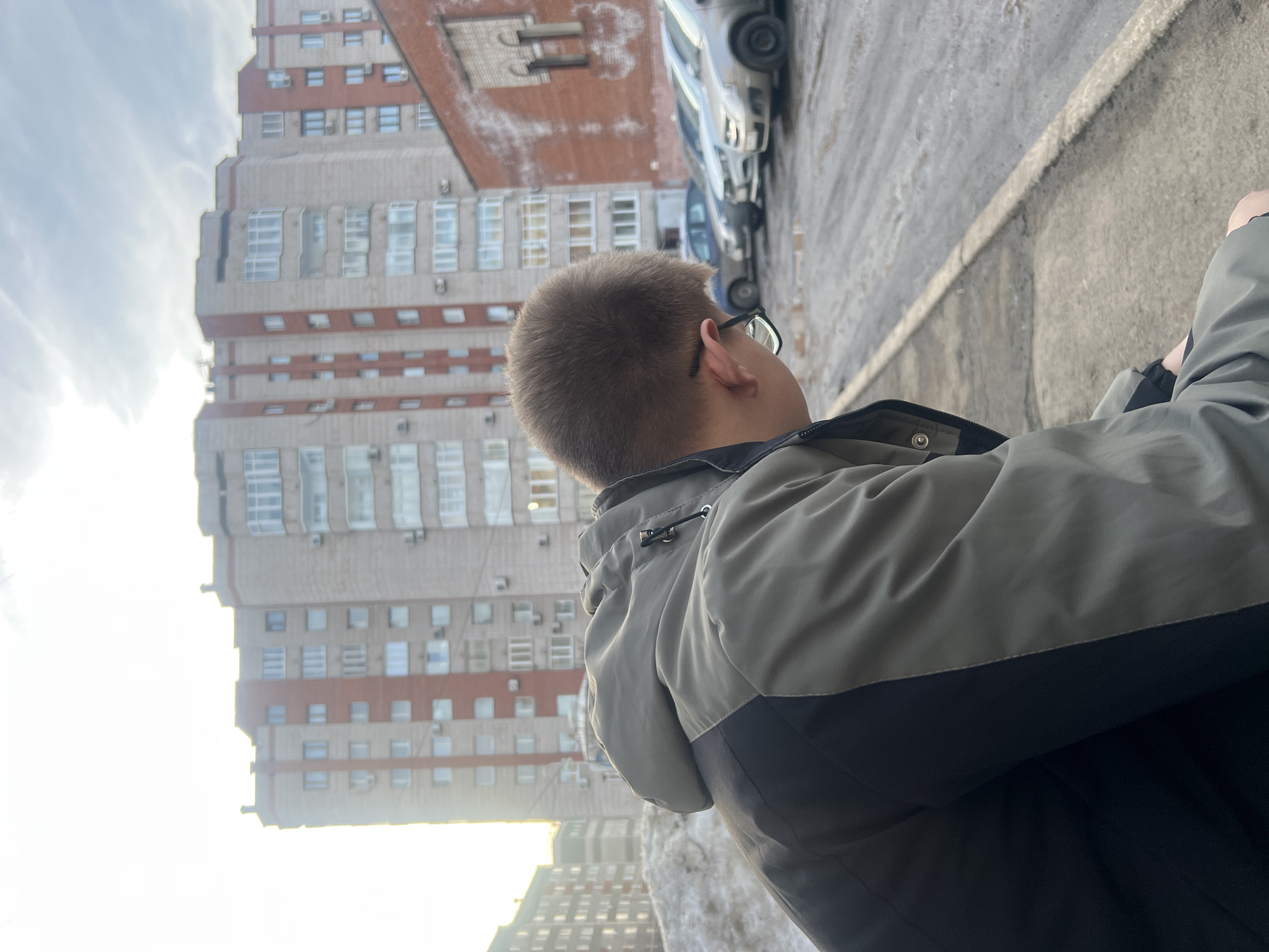 The image depicts an urban scene on a residential street within the city of Izhevsk, Russia, likely during late winter or early spring due to the presence of snow and wet conditions.
In the immediate foreground, facing away from the viewer and angled slightly to the right, is an individual from the chest up. The person has short, light brown hair and wears glasses. They are clad in a two-tone jacket, primarily olive green, with black panels on the lower sleeves and sides. The individual is leaning forward, directing their gaze downward and to the right, outside the frame.
To the right of the individual, a concrete sidewalk transitions into a wet, dark asphalt surface, presumably a parking lot or street. This area is marked by several shallow puddles. A row of at least five passenger vehicles is parked on the asphalt, facing away from the viewer. These vehicles are generally light-colored, including silver, white, and a dark blue or grey, appearing to be compact sedans or hatchbacks.
In the mid-ground, a prominent multi-story residential building dominates the central background. This building features a light beige or grey facade with contrasting vertical reddish-brown sections. Numerous windows, many with white frames, are visible across its stories, alongside several external air conditioning units. To the far right, a section of a red brick building with distinctive arched white architectural detailing around a window is visible.
To the left of the central building, a large, dirty pile of melting snow is present. Behind this snow pile and to the far left of the frame, another multi-story residential building, slightly darker and less detailed due to bright backlighting, is visible. The sky is partly cloudy, with bright sunlight emanating from the left side of the frame, causing some overexposure in that area.