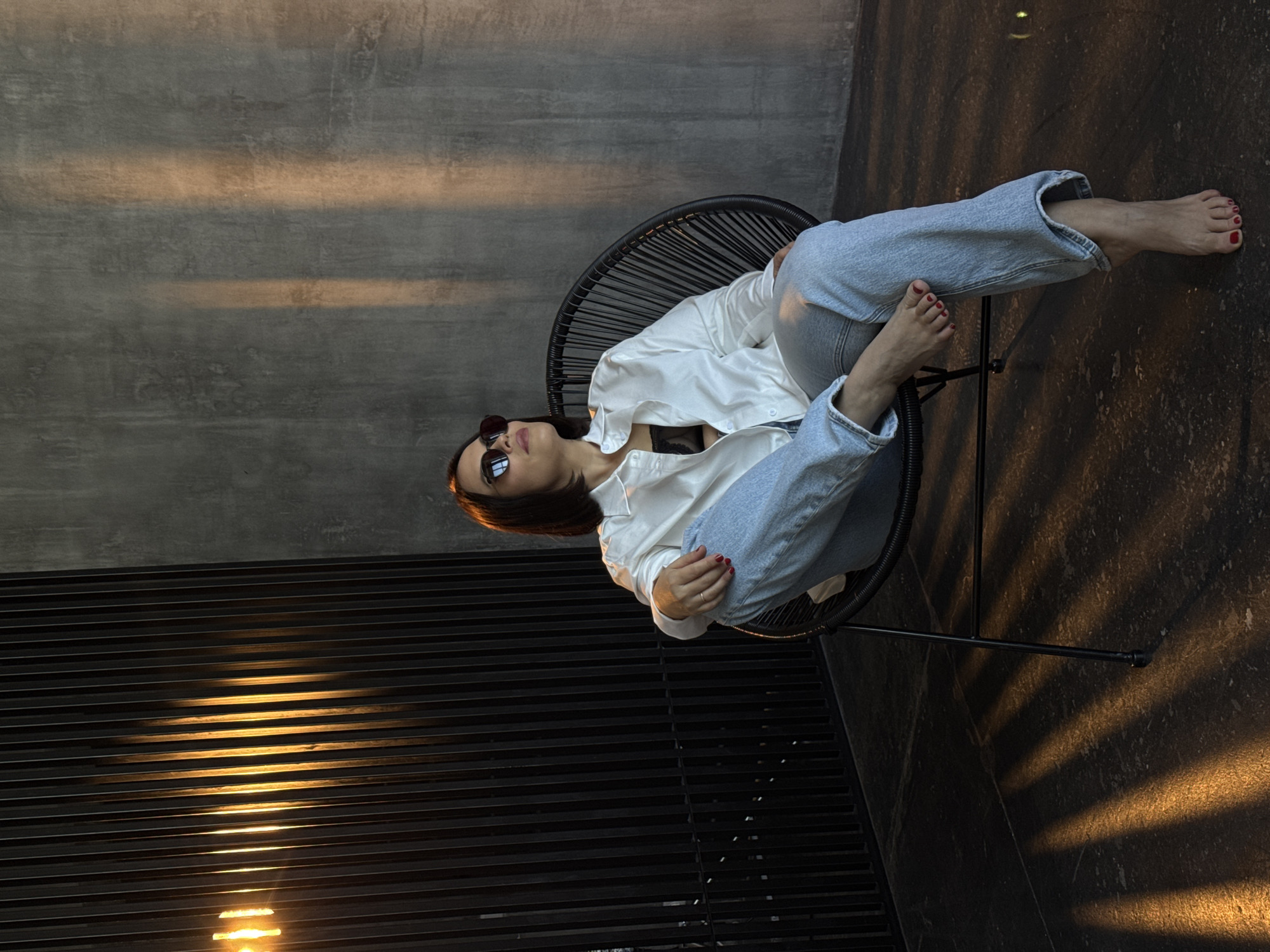 A woman with dark hair and sunglasses reclines in a black wire chair, wearing a white button-down shirt, a black bra, and light-wash denim jeans. Her feet are bare with red nail polish. The chair sits on a dark floor in a room with a gray wall. Light streams through horizontal slats, casting light and shadow patterns on the floor and wall. The location is Unknown, Unknown.