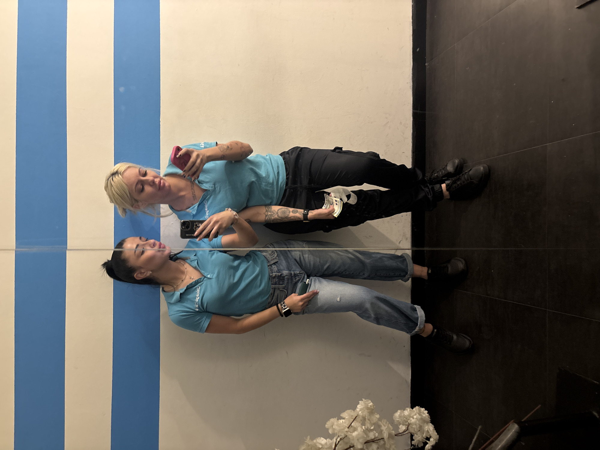 The image appears to be a mirror selfie taken by two women in what might be a commercial or public space.
**People:**
* Two women are visible. One has blonde hair and is wearing a light blue collared shirt and dark pants. She is holding a cell phone in one hand and currency in the other. The second woman has dark hair pulled back and is wearing a light blue collared shirt and jeans. She is holding a cell phone.
* Both women appear to be reflecting each other, indicating a mirror.
**Objects:**
* Cell phones, presumed to be used for taking the selfie.
* Currency (possibly euro or Bulgarian lev).
* Two different outfits worn by the women.
* A mirror reflecting the scene.
**Environment:**
* The background includes a wall with vertical blue and white stripes on one side and a plain white or light-colored wall.
* The floor is covered in black tiles. There are some objects or decorations in the bottom right corner, appearing to be faux white flowers.
**Actions/Interactions:**
* The women are taking a selfie in a mirror.
**Location:**
* While the image itself doesn't reveal the location, it is indicated to be in Plovdiv, Bulgaria. Without further details, it's impossible to pinpoint the specific location within the city.
* The context (commercial or public space) can't be deduced with certainty but is likely.