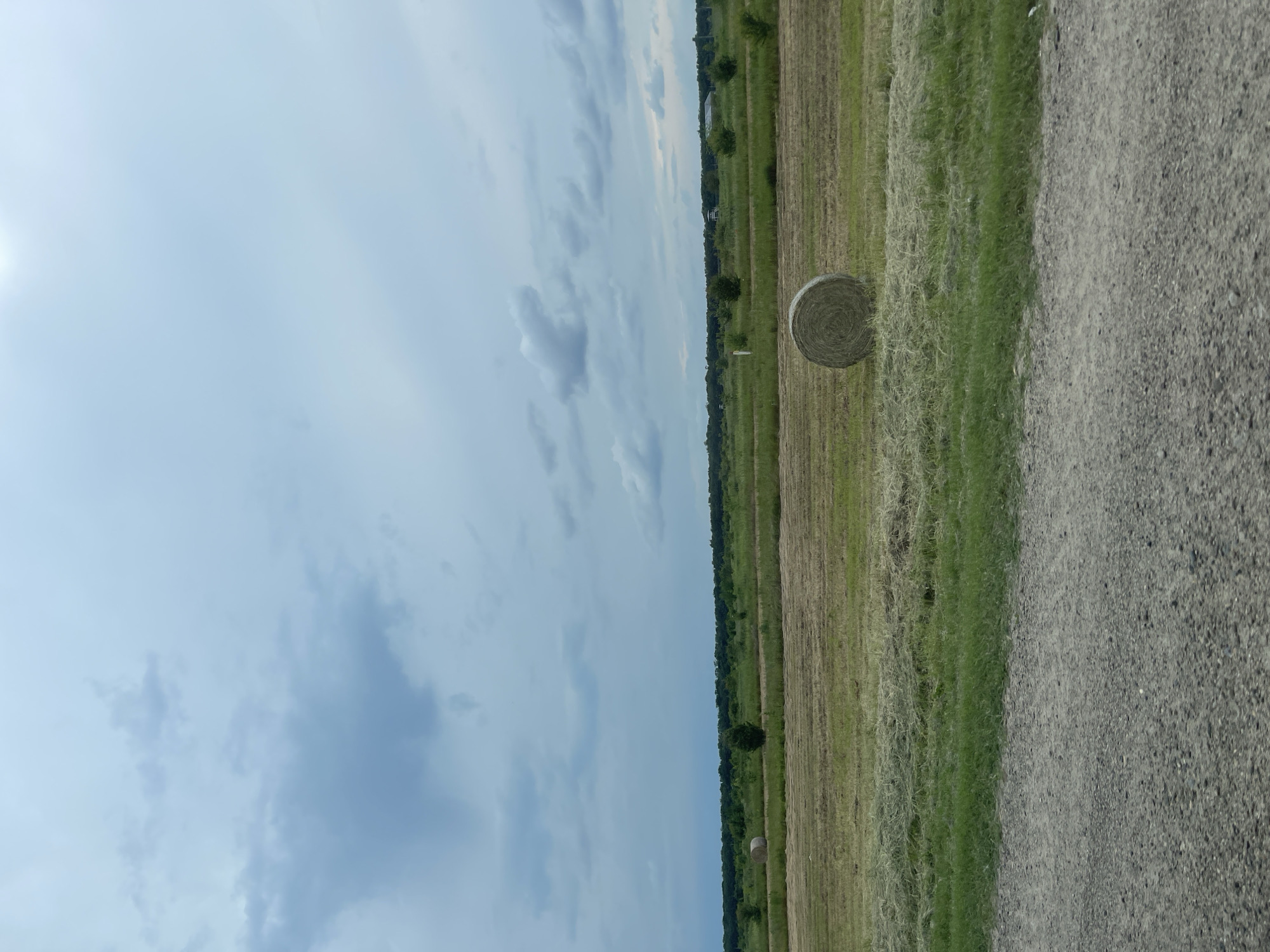 The image depicts a rural landscape with a hay bale in a field. The scene is oriented vertically, making it appear as if the viewer is looking upwards at the sky or downwards at the ground.
**Subjects:**
The primary subjects are a single, round hay bale and a field of cut grass. In the background, a line of trees marks the edge of the field, and beyond that, a strip of greenery stretches towards the horizon. A gravel road or path is visible on the right side of the frame. The sky occupies a significant portion of the image, filled with scattered clouds.
**Activity or Event:**
The scene suggests a harvested field, indicating that the hay has been baled. The presence of the hay bale implies an agricultural activity.
**Notable Details:**
* **Time of Day:** The lighting, with a bright area on the left and the overall illumination, suggests it is daytime, possibly late morning or early afternoon.
* **Weather:** The sky is partly cloudy, with some areas of brighter sky and some darker, heavier clouds. This indicates a mix of sunshine and cloud cover.
* **Location Cues:** The vast, open field, the presence of hay bales, and the tree line are typical of a rural or agricultural setting.
* **Emotions:** The image conveys a sense of peacefulness and the vastness of nature.
**Visible Text:**
There is no visible text in the image.