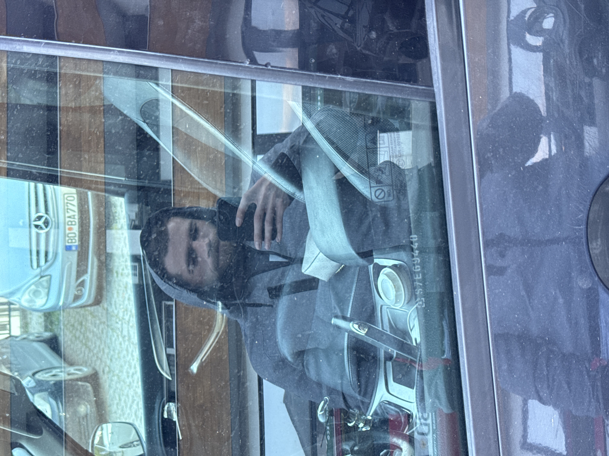 The image captures a reflection of a person and their surroundings in a car window, rotated ninety degrees clockwise. The primary subject is a male figure, seen from the chest up, wearing a hooded top. He is holding a smartphone in his right hand, seemingly taking a picture of his reflection or the car's interior through the window.
Inside the car, visible through the reflection, are parts of the dashboard and a seatbelt. The dashboard shows a steering column and some circular controls. A light-colored seatbelt stretches across the view, connected to the upper frame. On the windshield, obscured by dirt and reflections, the text "57E69440" is visible, likely a vehicle identification number or part number. Another sticker with partially readable text, possibly including "E11 43R-001021", is also present.
The car window itself appears dusty or dirty, contributing to a slightly hazy view. Numerous reflections are visible, indicating bright, possibly sunny, daytime conditions. In the upper left portion of the image (as oriented in the original file), the reflection of a light blue Mercedes-Benz vehicle is prominent, with a license plate "BD BA770". The "BD" prefix indicates Budva, Montenegro, confirming the provided location context. Reflections of other buildings or structures with vertical wooden or striped patterns are also present in the background. Another car's rearview mirror and front fender are reflected towards the bottom of the image.