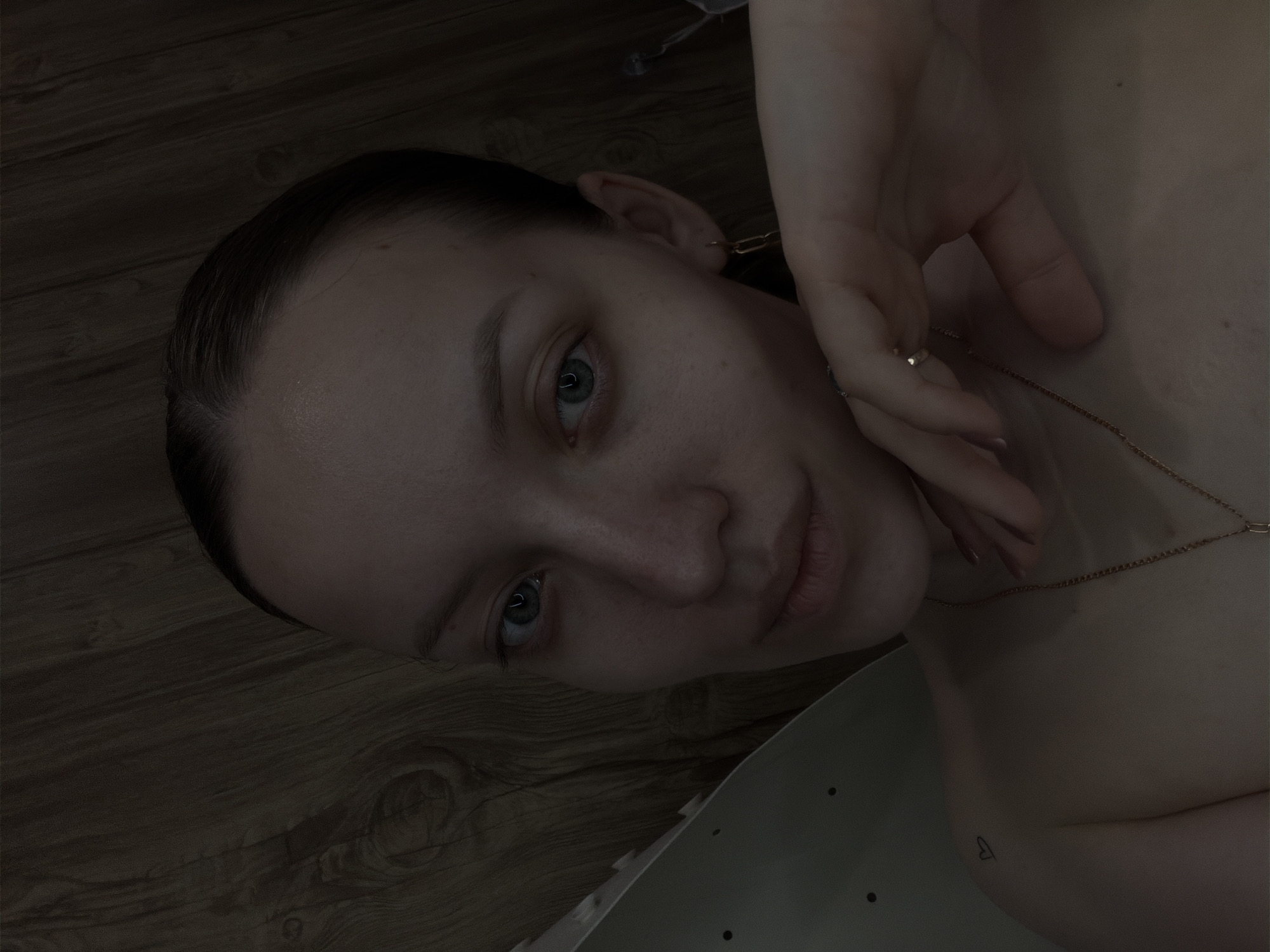 A close-up, dimly lit portrait shows a woman with light skin, blue eyes, and dark hair pulled back. Her face is angled towards the viewer, and her hand, adorned with a ring, is positioned near her face. She wears a thin gold necklace. A small heart tattoo is visible on her shoulder. The background includes what appears to be a wooden floor and part of a white surface. The location is unspecified.