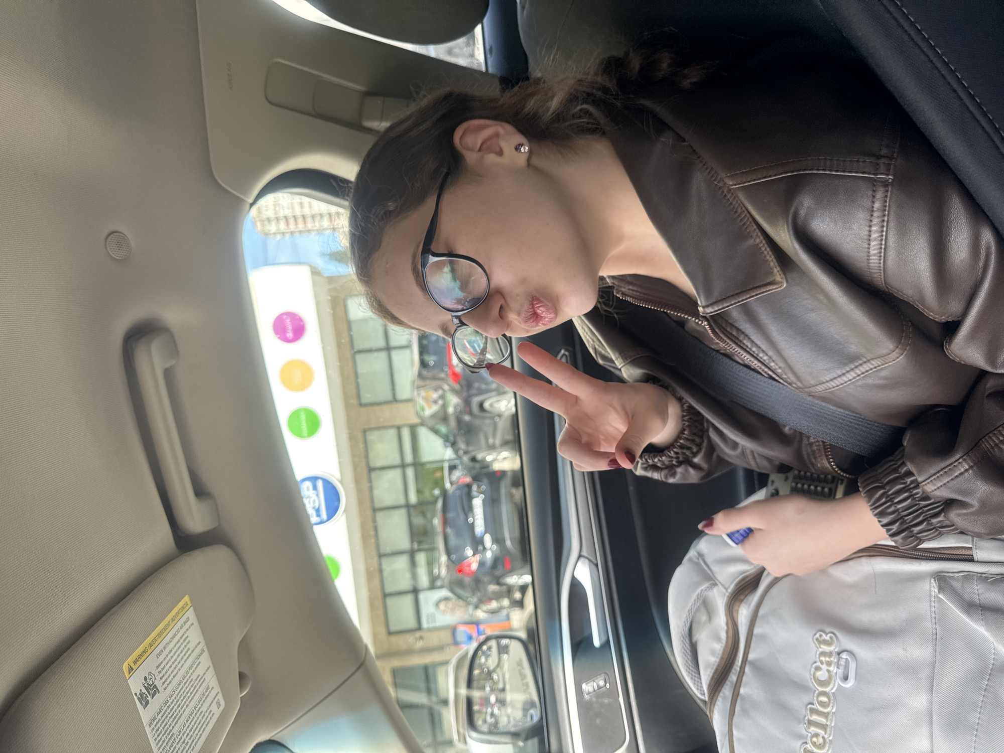 A young person is captured in a playful moment inside a car, likely in the passenger seat. They have long, dark hair, wear glasses, and are dressed in a brown leather jacket. The individual is making a "duck face" with their lips and holding up a peace sign with their right hand, suggesting a lighthearted mood. A small stud earring is visible in their ear.
The setting is daytime, with bright light coming from outside the car, indicating sunny or partly cloudy weather. Through the car window, an urban environment is visible with buildings and other vehicles parked or driving. Several colorful circular signs are seen outside, one of which has "PSP" printed on it. The interior of the car shows part of the ceiling, a sun visor with a warning label, and the passenger door panel.
Visible text on the sun visor includes "A WARNING/AVERTISSEMENT JADVERTENCIA," "EVEN WITH ADVANCED AIR BAGS," "MEME AVEC DES SACS GONFLABLES PERFECTIONNES," and a partial "LUSIVE CON LAS NUEVAS BOLSAS DE ARE." A light-colored backpack is resting on the seat next to the person, clearly displaying the word "HelloCat" (partially visible as "ellocat" in the crop) on its front. The scene is situated in Tbilisi, Georgia.