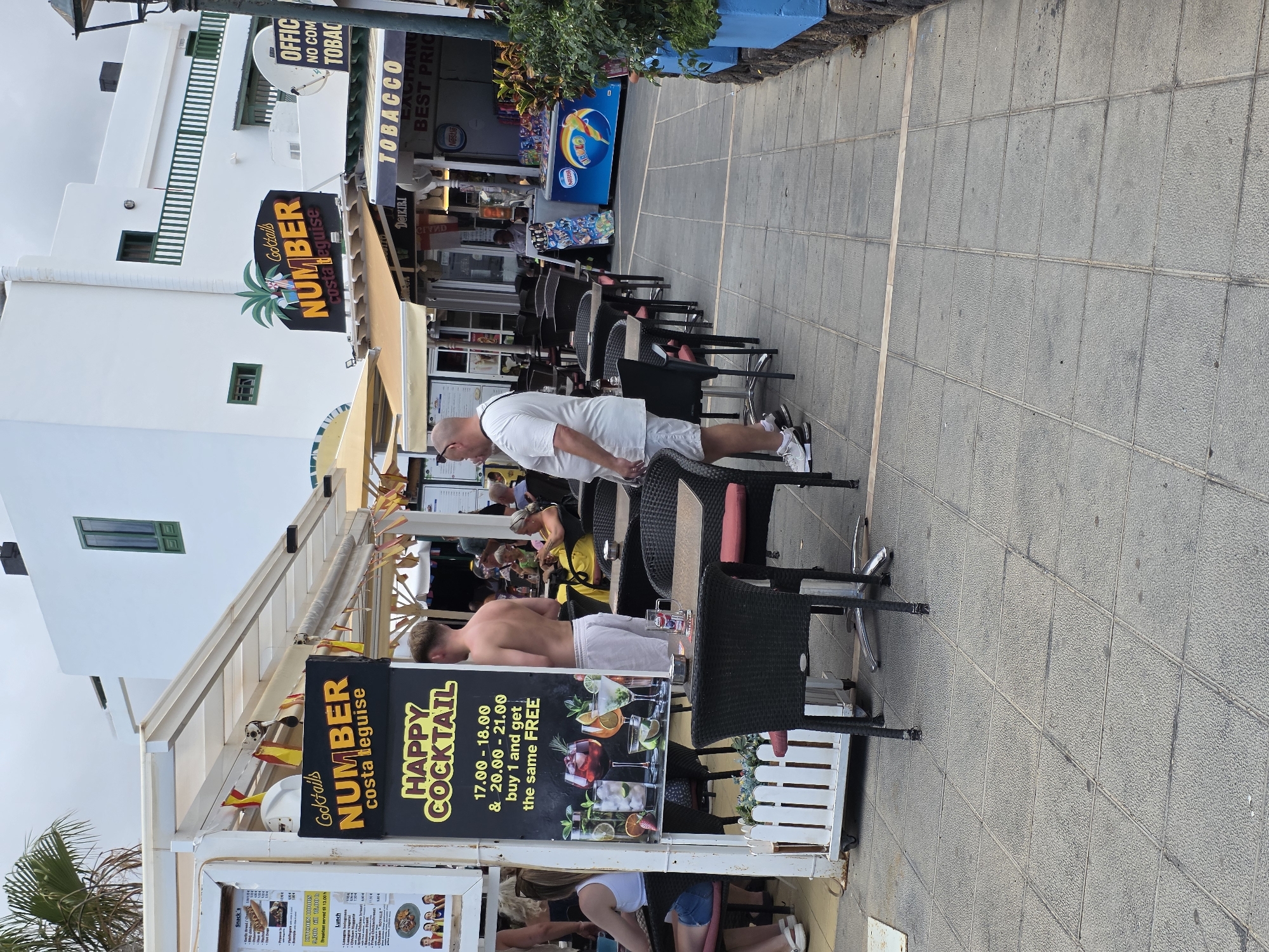 This is a photograph taken outdoors in Costa Teguise, Spain, likely during daylight hours given the brightness. The scene depicts a street-level view of a cafe or bar with outdoor seating.
Several people are visible, including a shirtless man in white shorts standing near a table, and other patrons seated or standing in the background. The cafe features black chairs and tables set up on a paved patio. A prominent sign for "NUMBER cocktails costa teguise" is visible, advertising a "HAPPY COCKTAIL" special with specific hours and a "buy 1 and get the same FREE" offer. Another sign indicates "TOBACCO EXCHANGE BEST PRIC". There are also signs for "Snack's" and "Lunch" with menu items listed. A refrigerator displaying ice cream is also present. In the background, white buildings with green accents line the street, and some vegetation is visible. The overall atmosphere appears casual and relaxed, consistent with a holiday destination.
