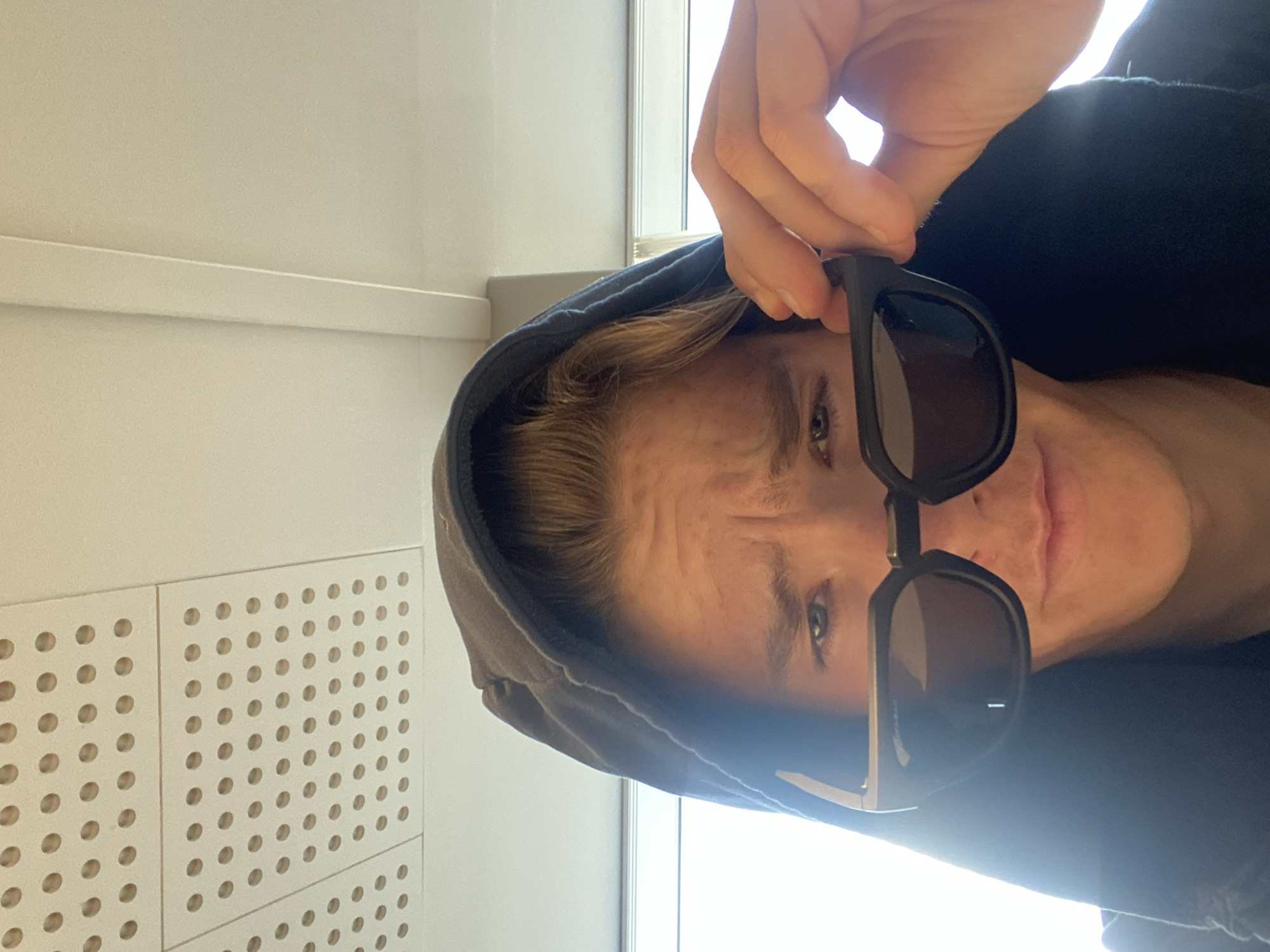 A person is holding a pair of sunglasses down and in front of their face, partially obscuring their eyes. The person is wearing a gray hooded sweatshirt. The background includes a white wall with a grid of circular perforations and a window with a white frame. The photo was taken indoors. There is no indication this photo was taken in Kastrup, Denmark.