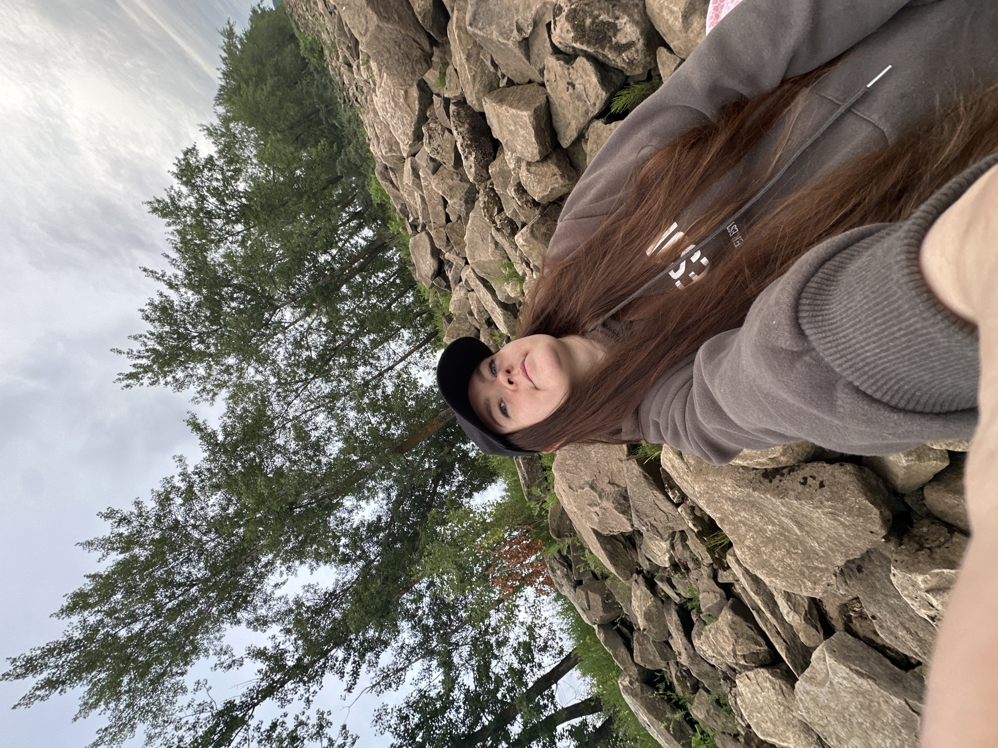 The image shows a young woman with long, reddish-brown hair wearing a gray hoodie and a black baseball cap, taking a selfie outdoors. She is positioned on a rocky incline, with large, gray boulders and some sparse green vegetation filling the foreground and midground. Behind her, a line of tall trees with green foliage stretches across the scene, their branches reaching towards a cloudy, overcast sky. The ground is uneven and covered in rocks of varying sizes, suggesting a natural or semi-natural environment. The overall impression is one of a casual outdoor setting. Given the context provided, this scene is located in Chaikovsky, Russia.
