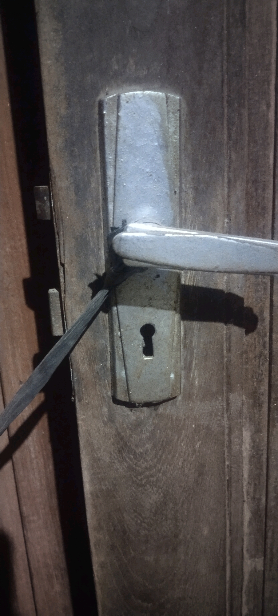 The image shows a close-up, eye-level view of a wooden door in Daleman, Indonesia. The door features a metal door handle mechanism with a keyhole. A black cable or wire is wrapped around the handle and appears to be securing it or potentially being used to tamper with the lock. The wood grain is visible on the door, and there are some signs of wear and tear on both the door and the metal hardware. The lighting suggests an indoor environment or an area with artificial light, casting shadows on the door. No people are visible in the frame.